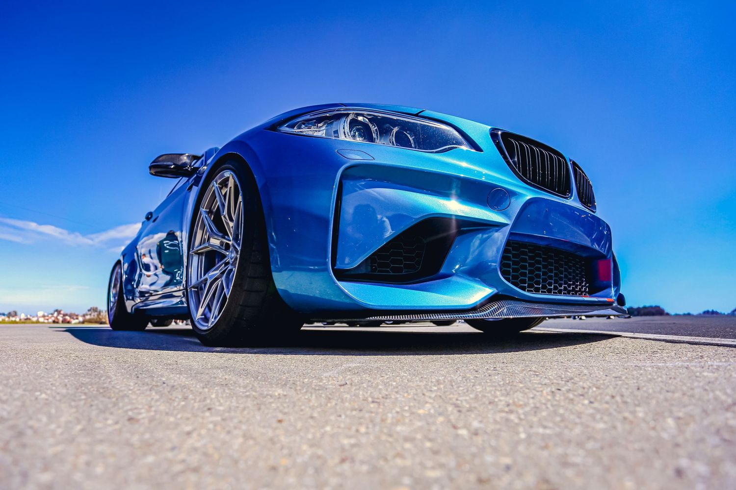 Low ground-level perspective of a blue BMW M2 with wide body kit under a vivid blue sky.
