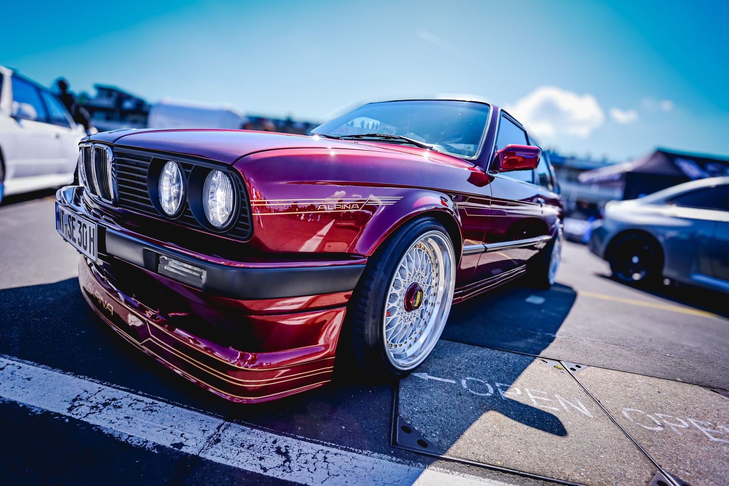 A heavily modified deep red BMW E30 with BBS-style wheels sits low at an outdoor car show.