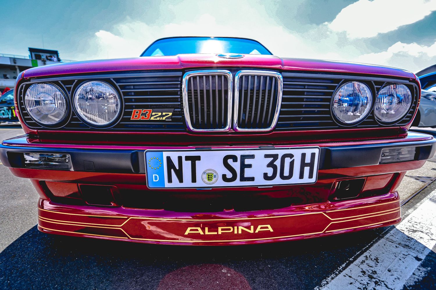 Red BMW Alpina E30 with NT SE 30 H license plate and quad headlights at a car meet.