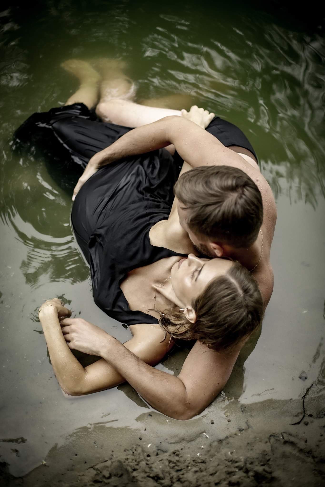 A couple in black clothing romantically embracing while lying in shallow water.