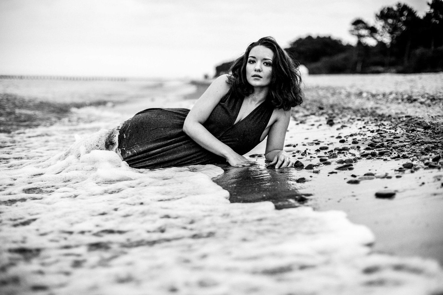 Black and white photo of a woman in a dark dress posing in shallow ocean waves on a beach.