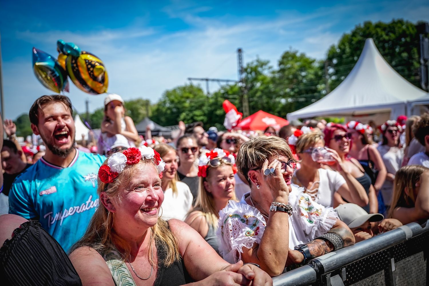 Excited crowd enjoying an outdoor music festival on a sunny day, with women wearing floral crowns in the foreground.