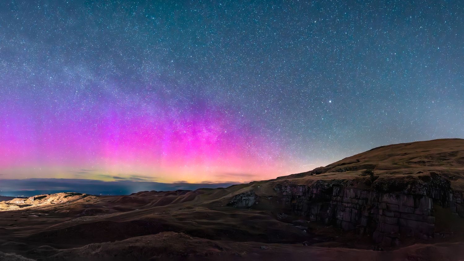 A vibrant display of the Northern Lights (aurora borealis) in pink and green hues over a dark, silhouetted landscape, Brecon Beacons, Wales
