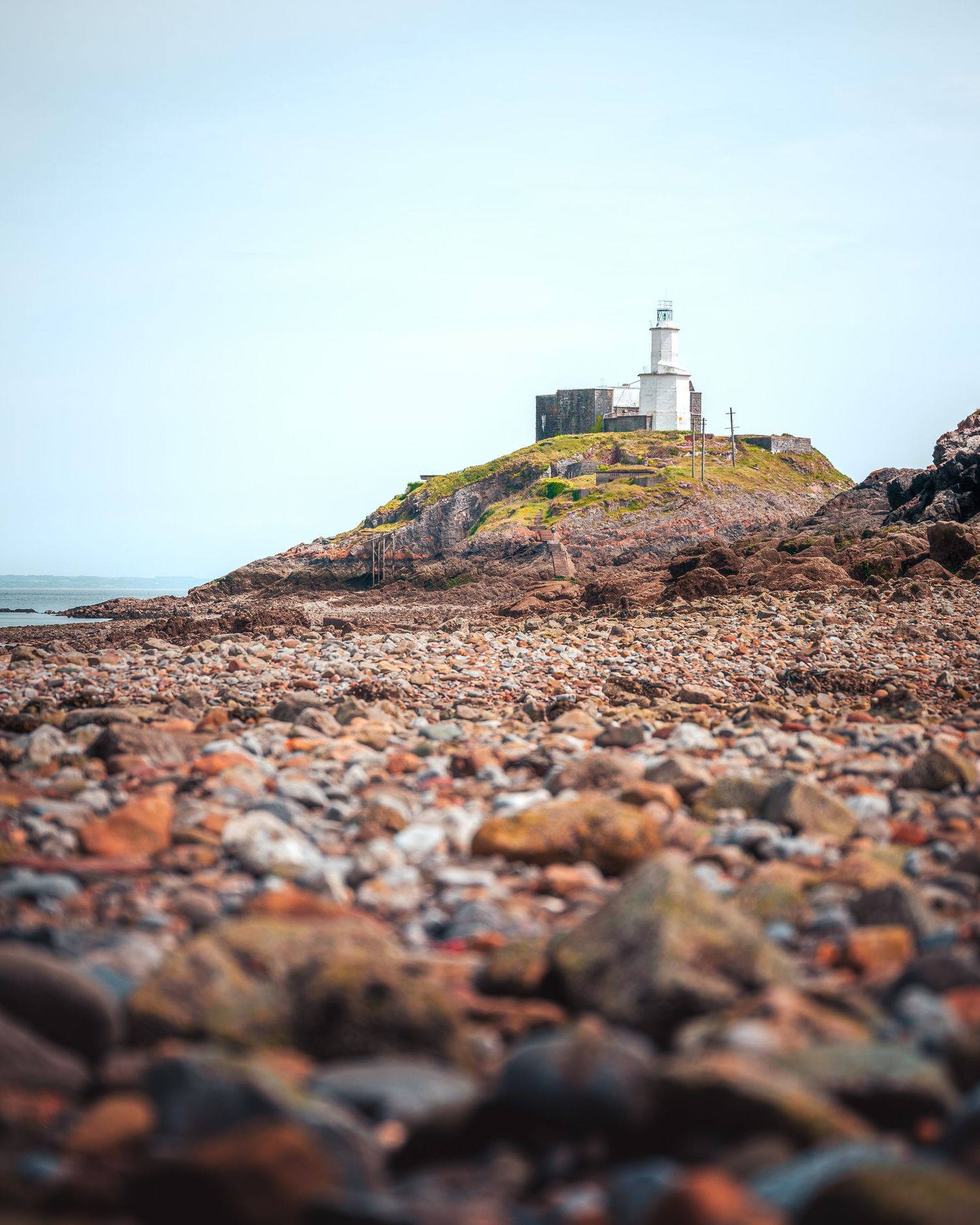 A rocky beach with a small, white lighthouse on a hill in the distance, Mumbles, Wales