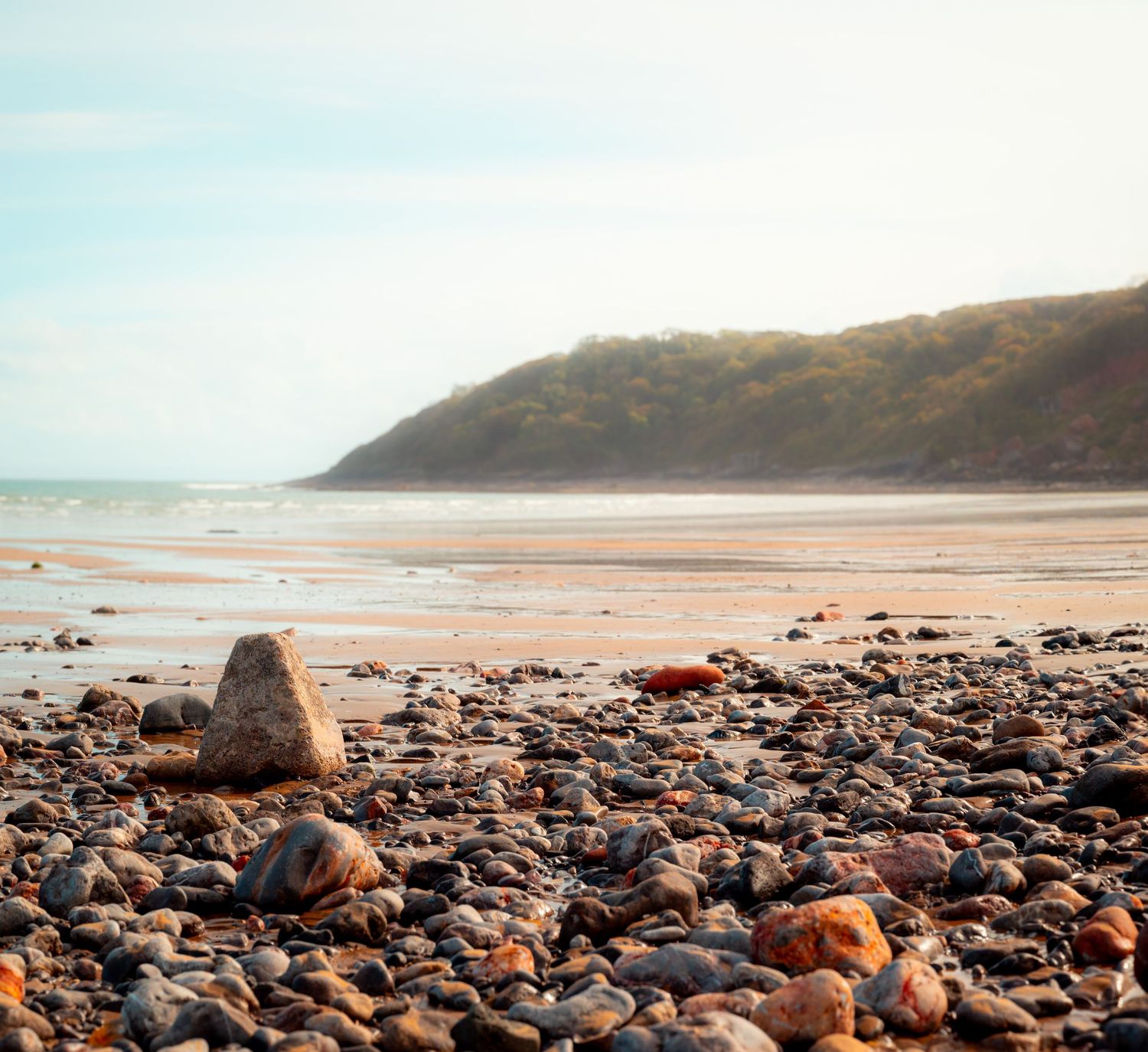 A rocky beach with a large, smooth stone in the foreground and a grassy hill in the distance under a bright sky, Oxwich Bay, Wales