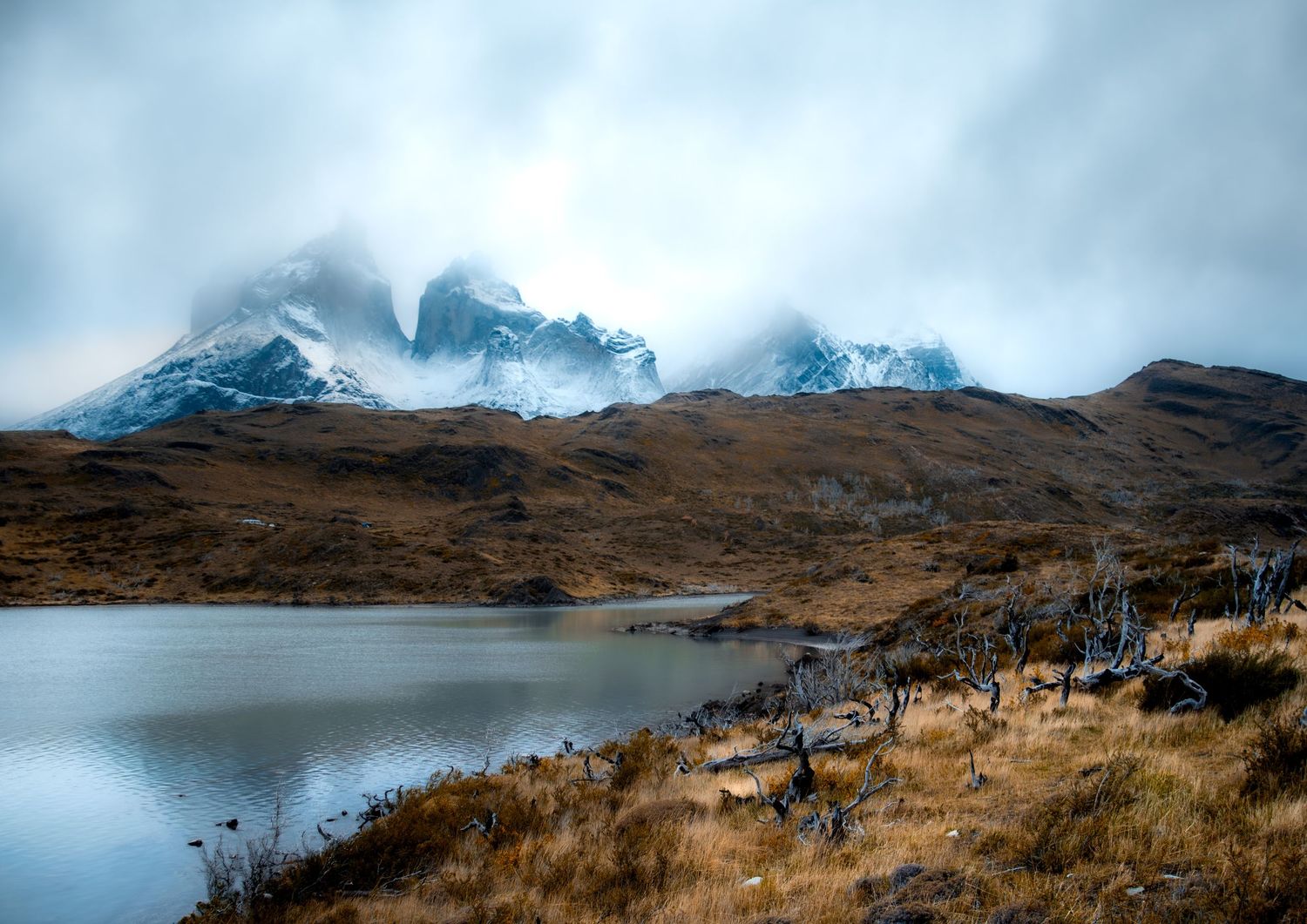 A calm lake reflecting snow-capped mountains under a cloudy sky, in Torres del Paine, Chile.