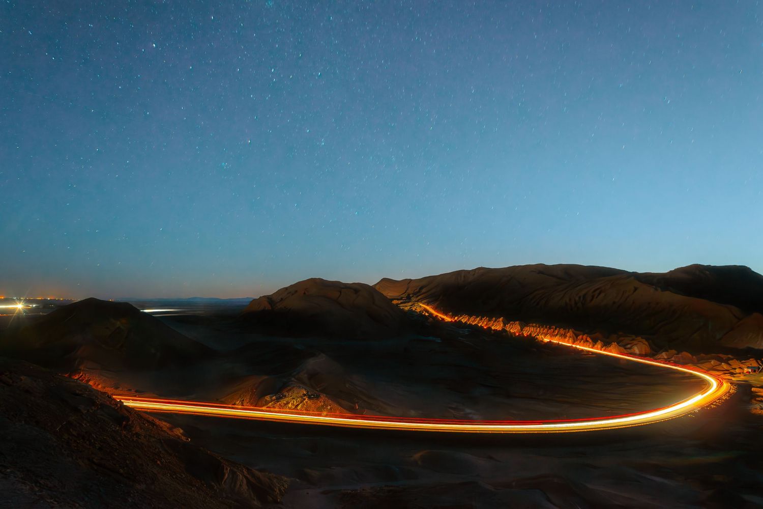 A long exposure photo of car headlights creating a winding trail of light across a dark desert landscape at dusk.