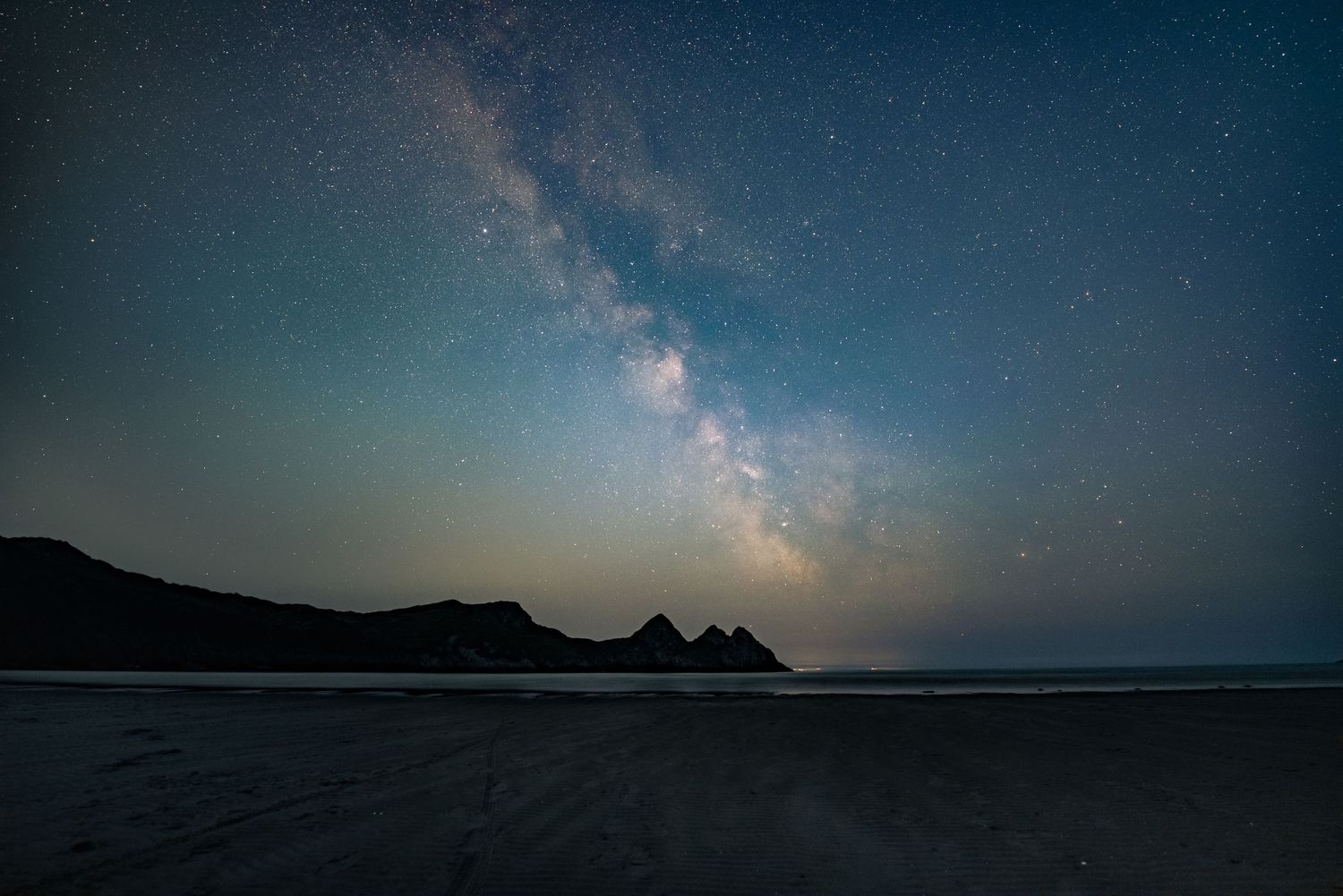 A starry night sky with the Milky Way visible over a dark, silhouetted landscape and a calm body of water.