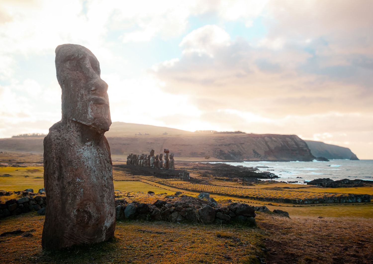 A large stone statue (Moai) standing on a grassy hill with the ocean and a cloudy sky in the background and another set of Moai.