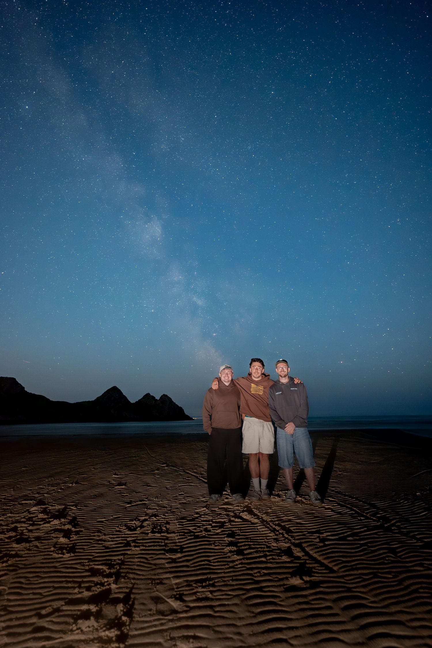 Three people standing close together on a dark beach at night, with the Milky Way visible in the sky above them.