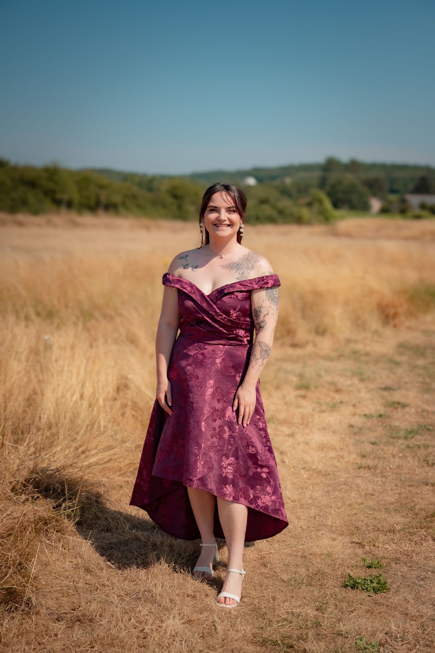 A woman in a long, dark red dress walking through a field of tall, dry grass under a blue sky.