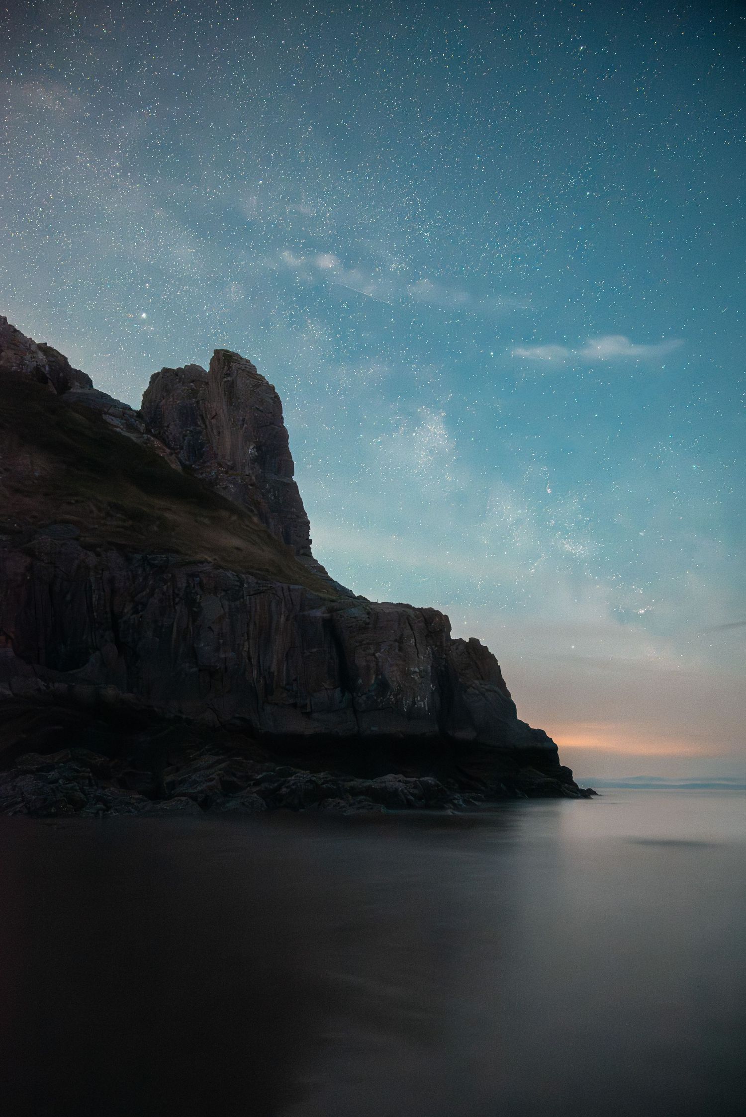 A dark, moody landscape with a large rock formation rising from a calm body of water under a cloudy sky, Tor Bay, Wales