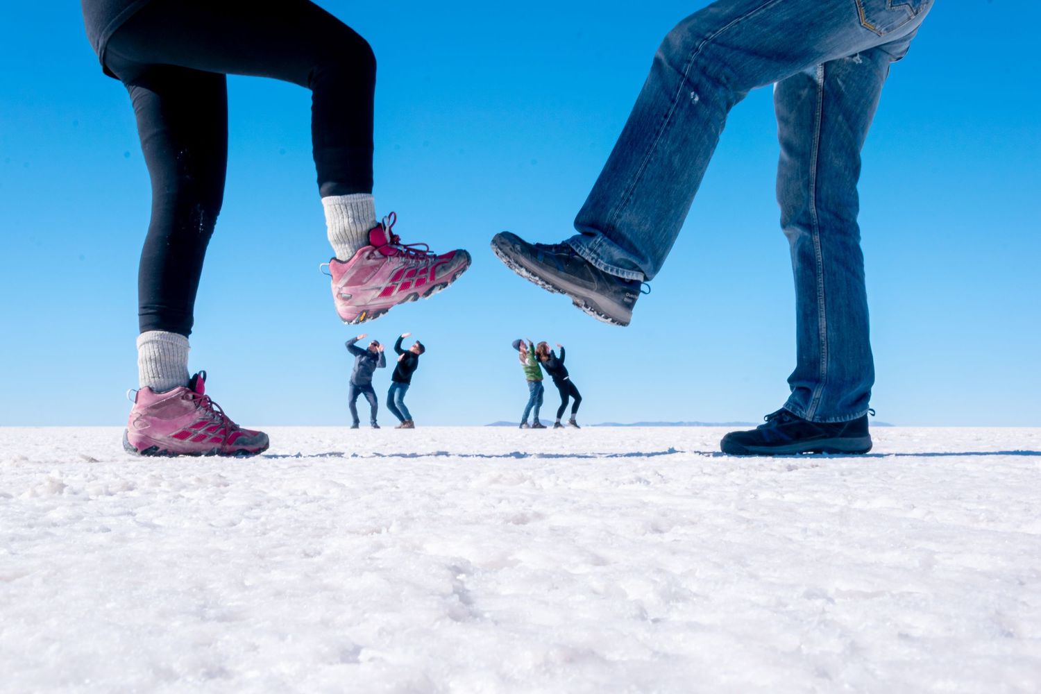 A forced perspective photo on a white salt flat, showing two people in the foreground appearing to step on a group of smaller people