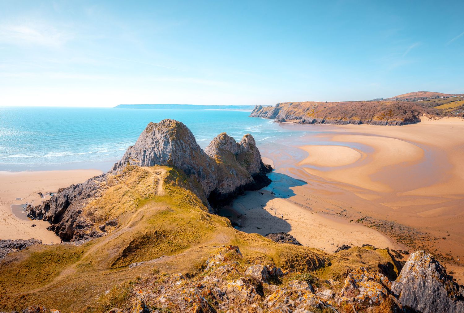 A wide, sandy beach with a large, rocky outcrop in the middle and a grassy headland in the background, Three Cliffs Bay, Wales