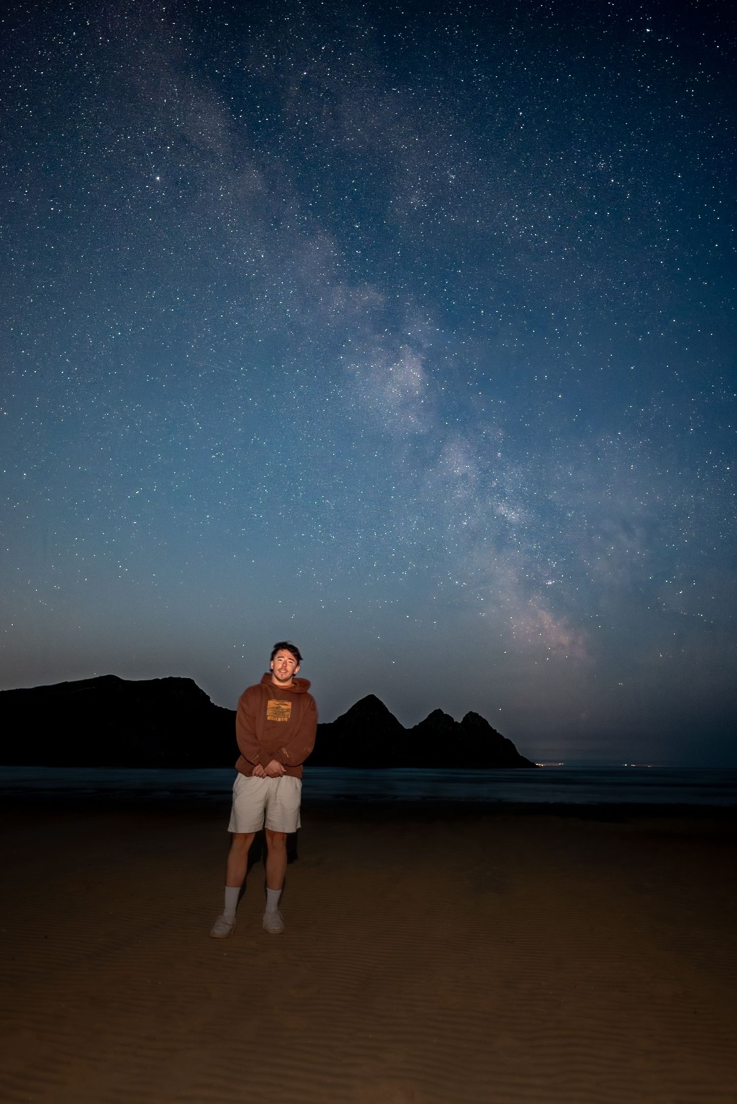 A person standing on a dark beach at night, looking up at a starry sky with the Milky Way clearly visible.