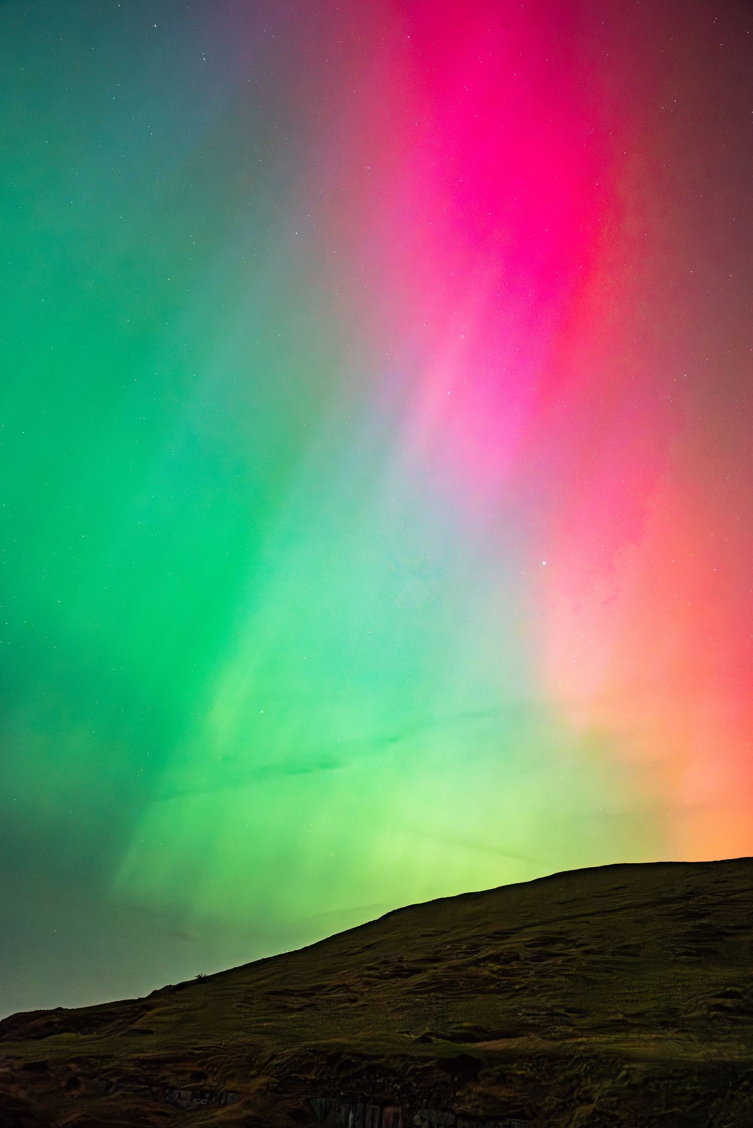 A vibrant display of the Northern Lights (aurora borealis) in green and pink hues across a dark night sky, Wales