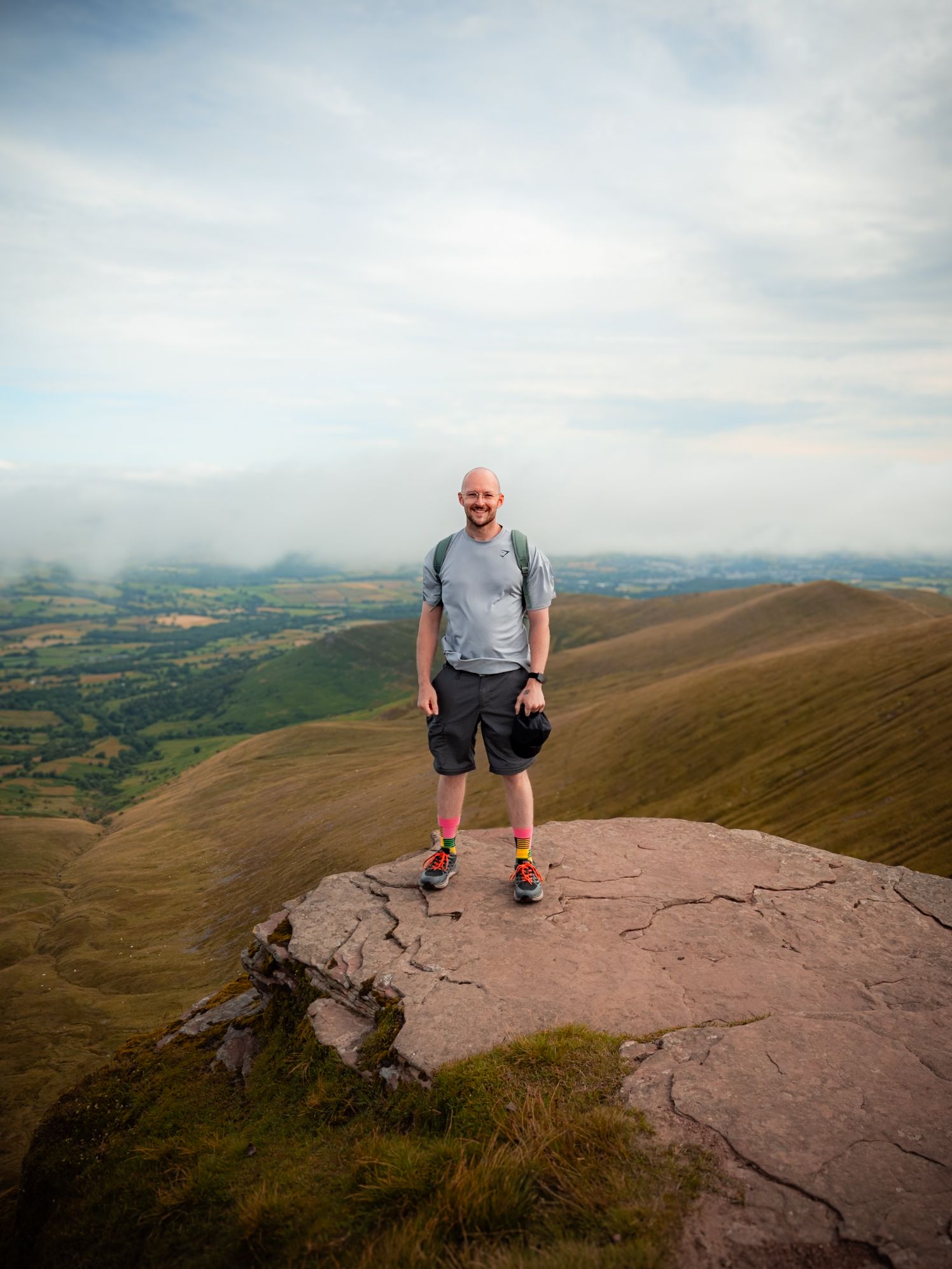 A man standing on a rocky outcrop overlooking a vast, hilly landscape under a cloudy sky.