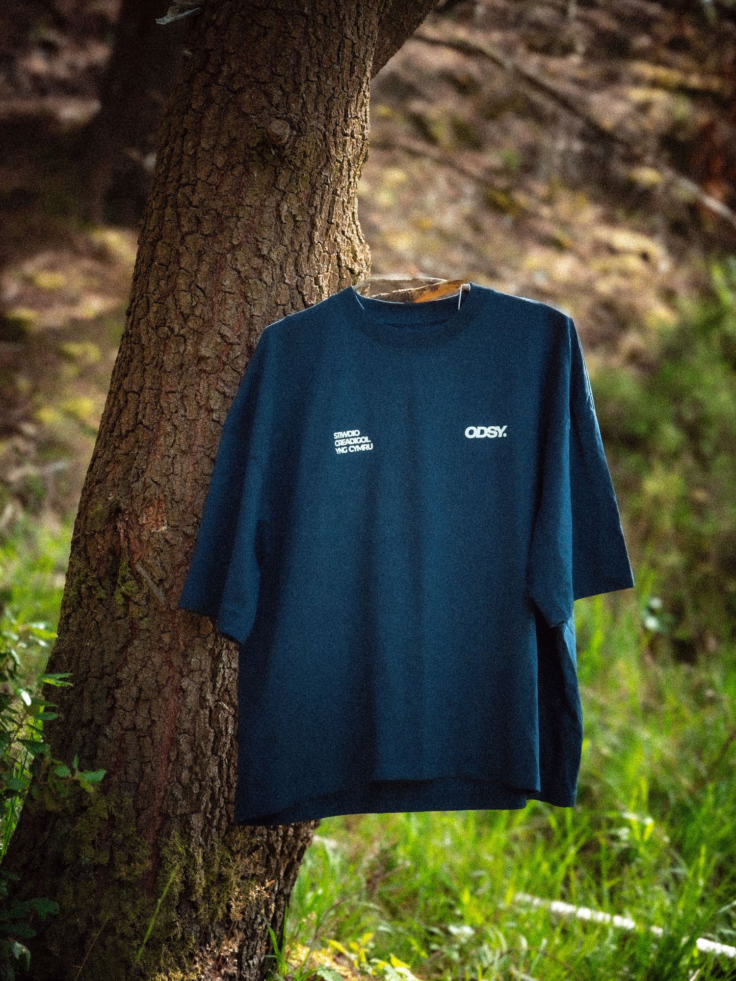 A dark blue t-shirt with a small white logo on the left chest area, hanging on a tree trunk in a sunlit forest.