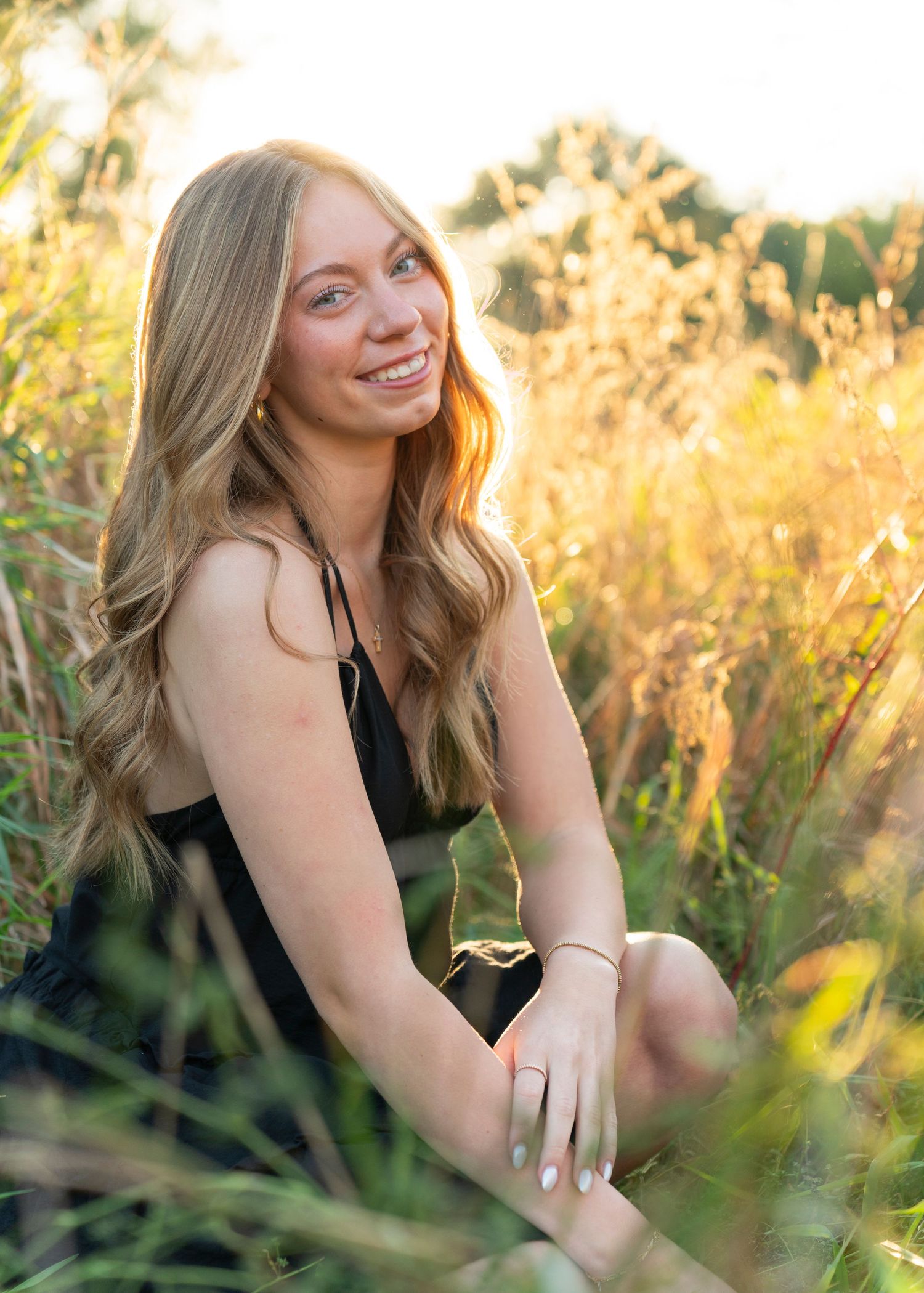 Smiling blonde woman in black dress sitting in golden sunlit tall grass field.