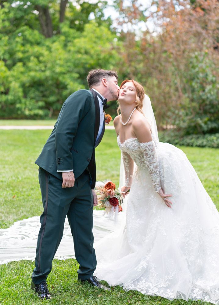 Bride and groom kissing on green lawn, bride holding bouquet in lace wedding gown.