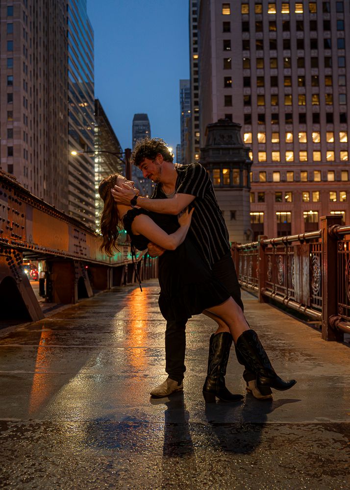 Couple shares romantic dip kiss on rain-soaked Chicago bridge at night with city lights glowing.