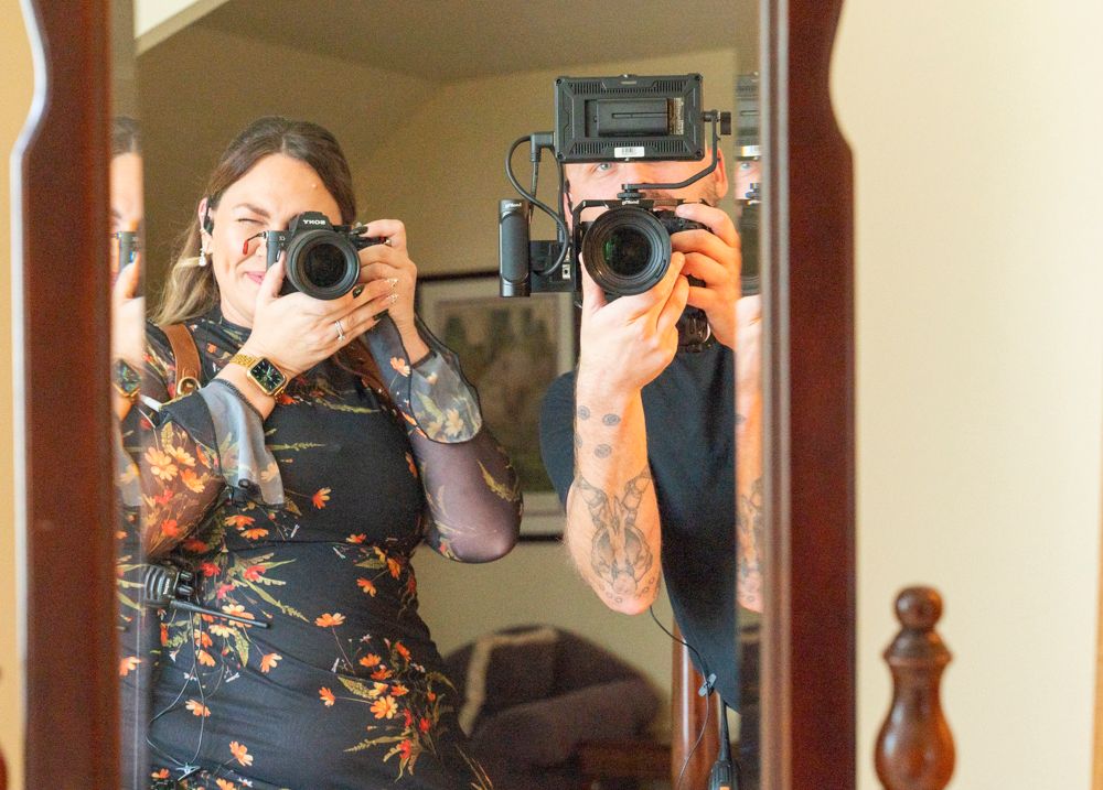 Two photographers taking a mirror selfie, one with tattoos holding a camera.