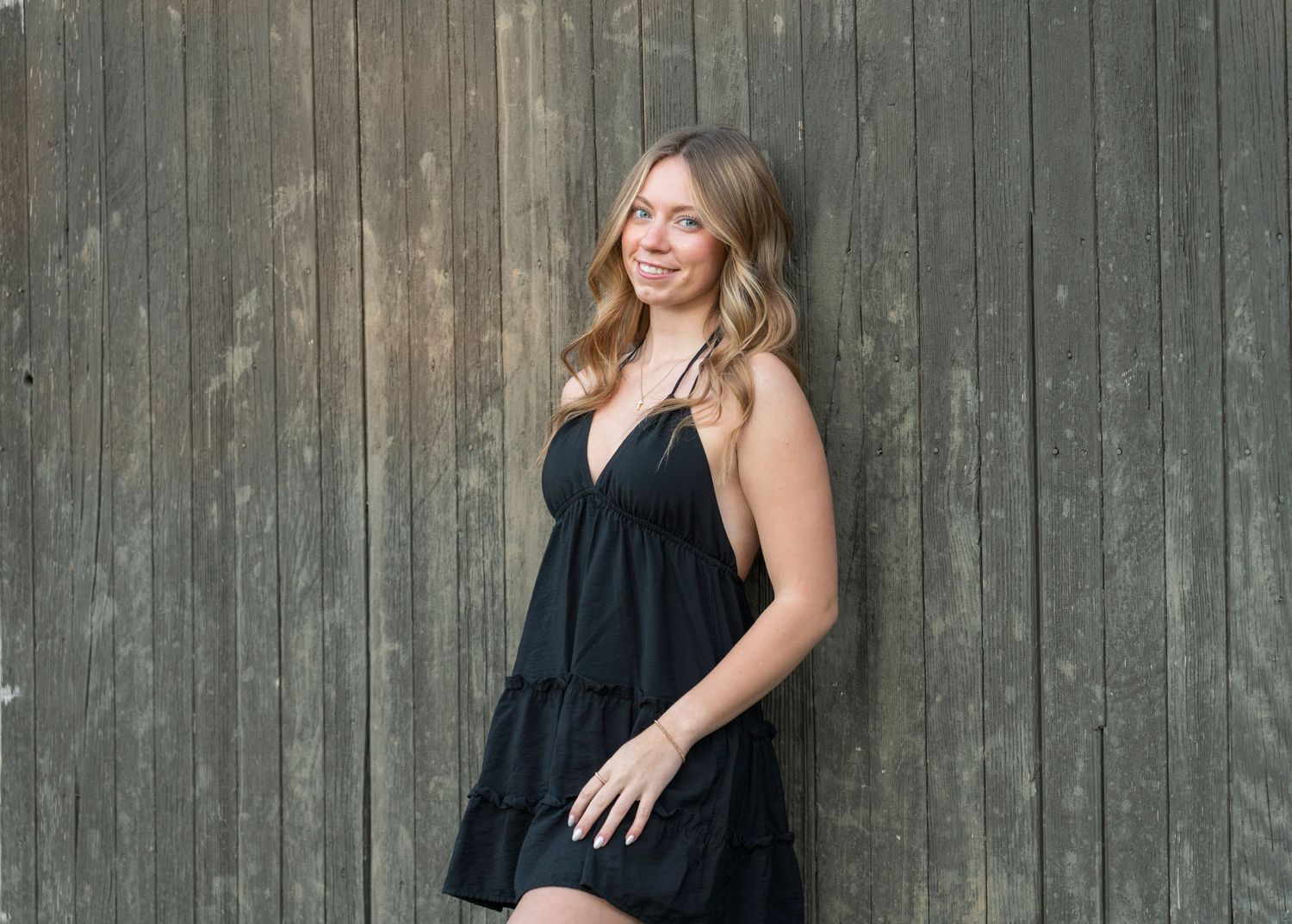 Smiling young woman in black sundress posing against a rustic wooden wall.