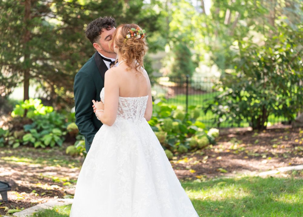 Groom in green suit kissing bride in white lace gown in a lush garden setting.