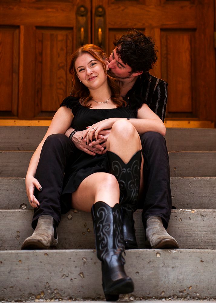 Couple in black outfits sit on stone steps, woman in cowboy boots smiling as man kisses her cheek.