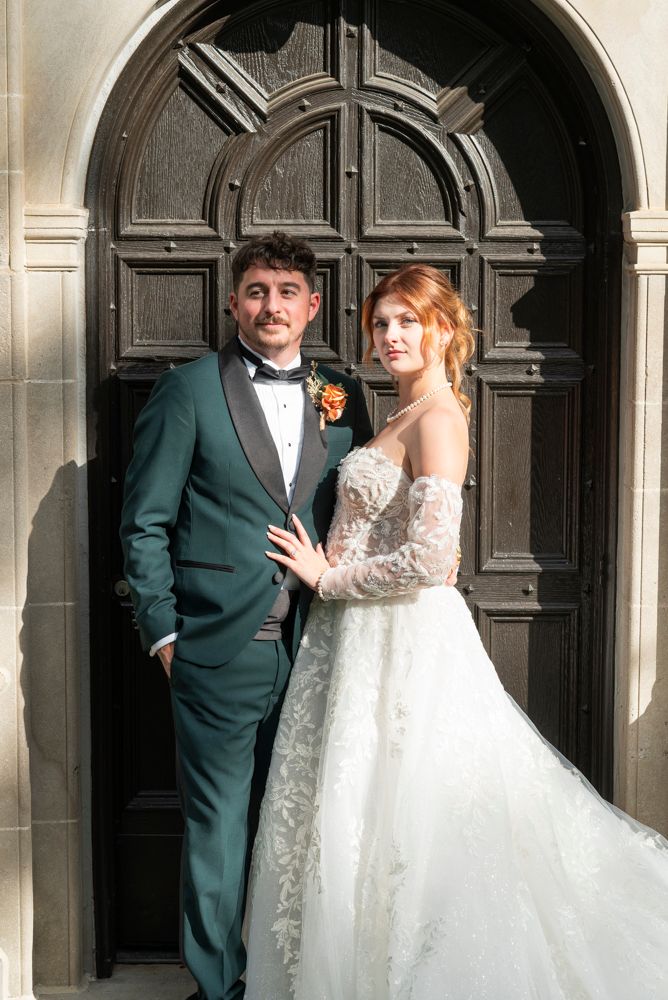 Bride in lace gown and groom in green tuxedo pose before ornate wooden church doors on wedding day.