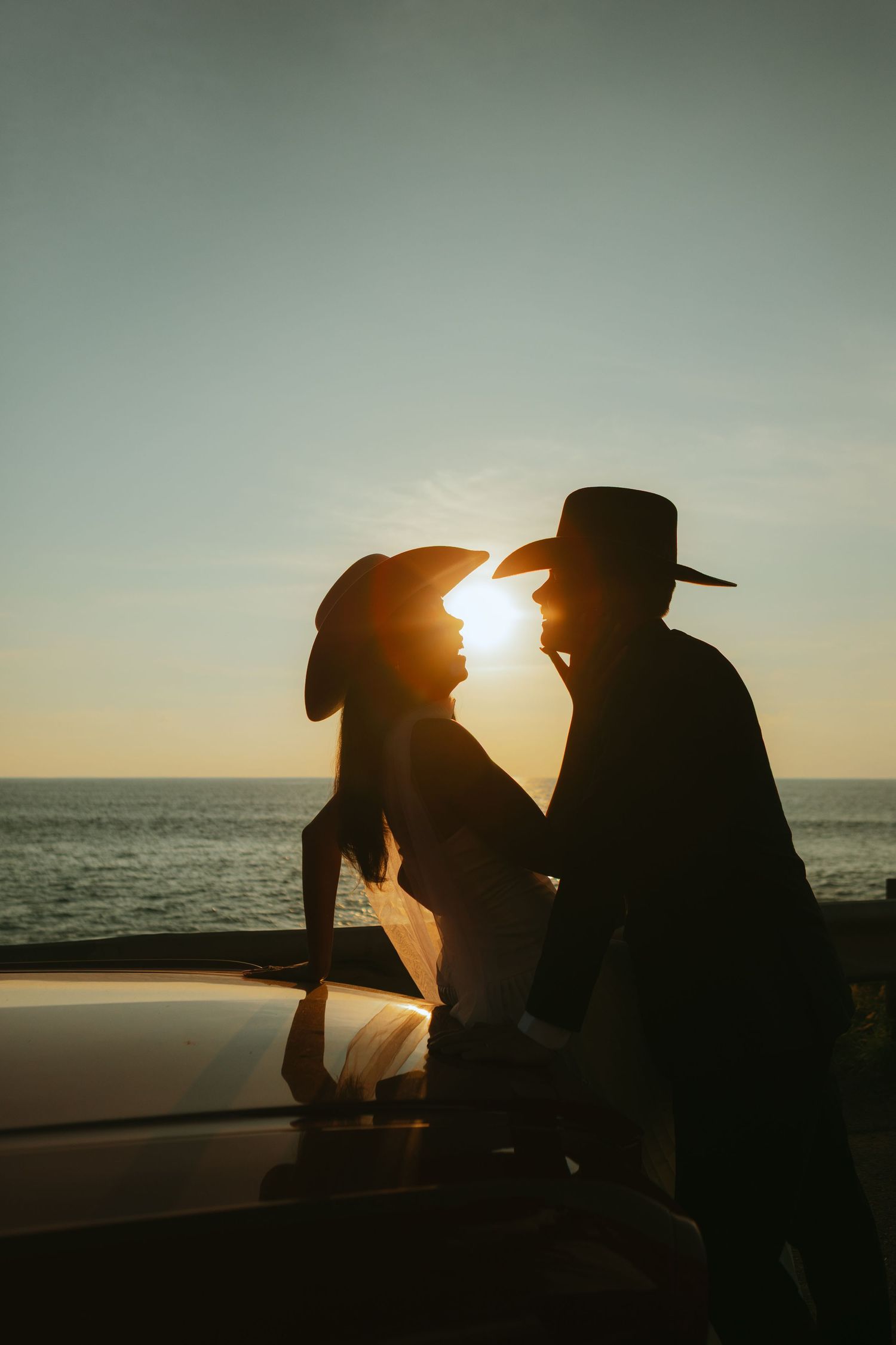 Silhouette of a romantic couple in wide-brim hats sharing a sunset kiss by the ocean on a car hood.