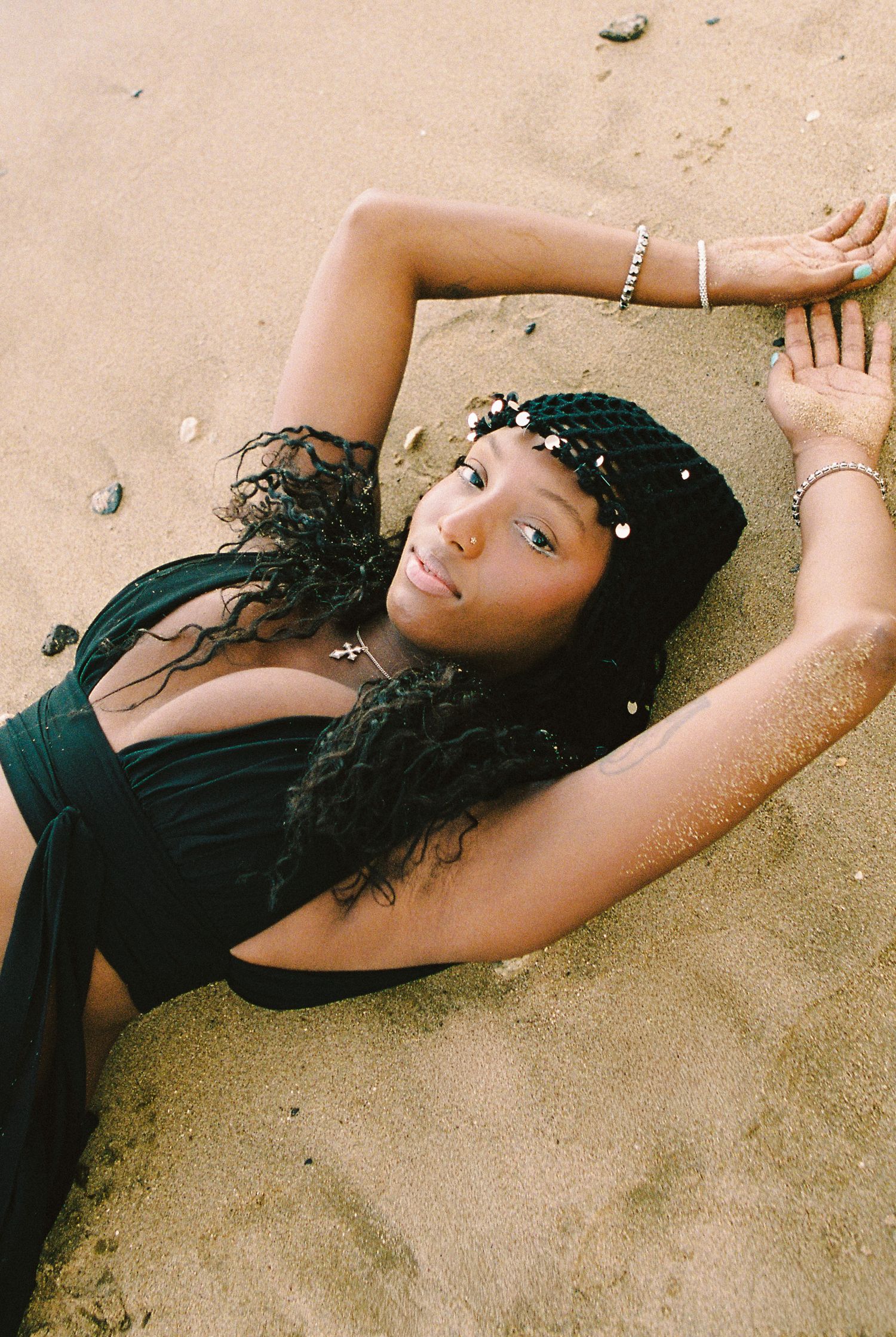 Young Black woman in black dress lying on sandy beach with arms raised, wearing jewelry.