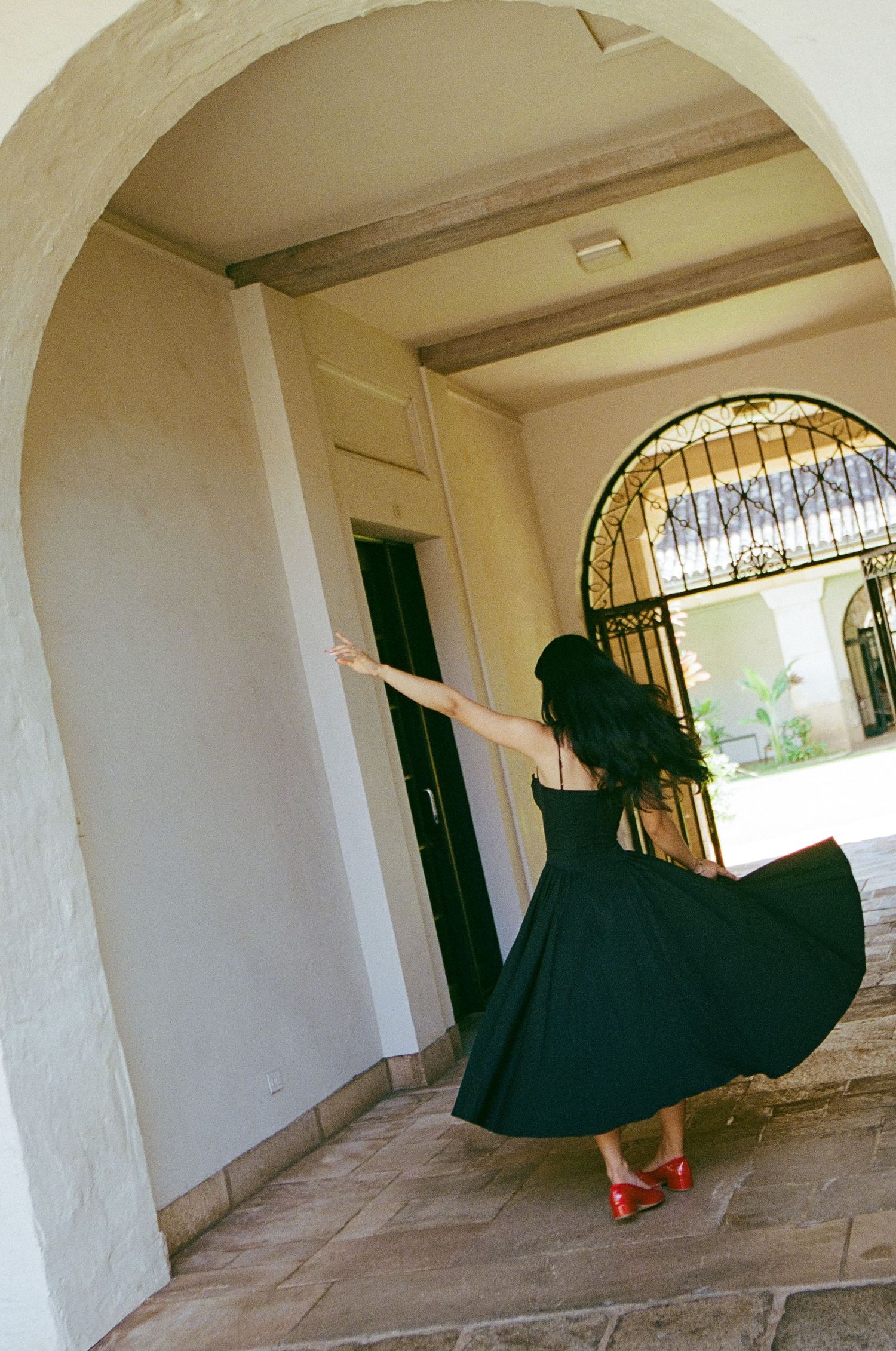 Woman in black dress and red heels twirling under an arched walkway near an ornate iron gate.