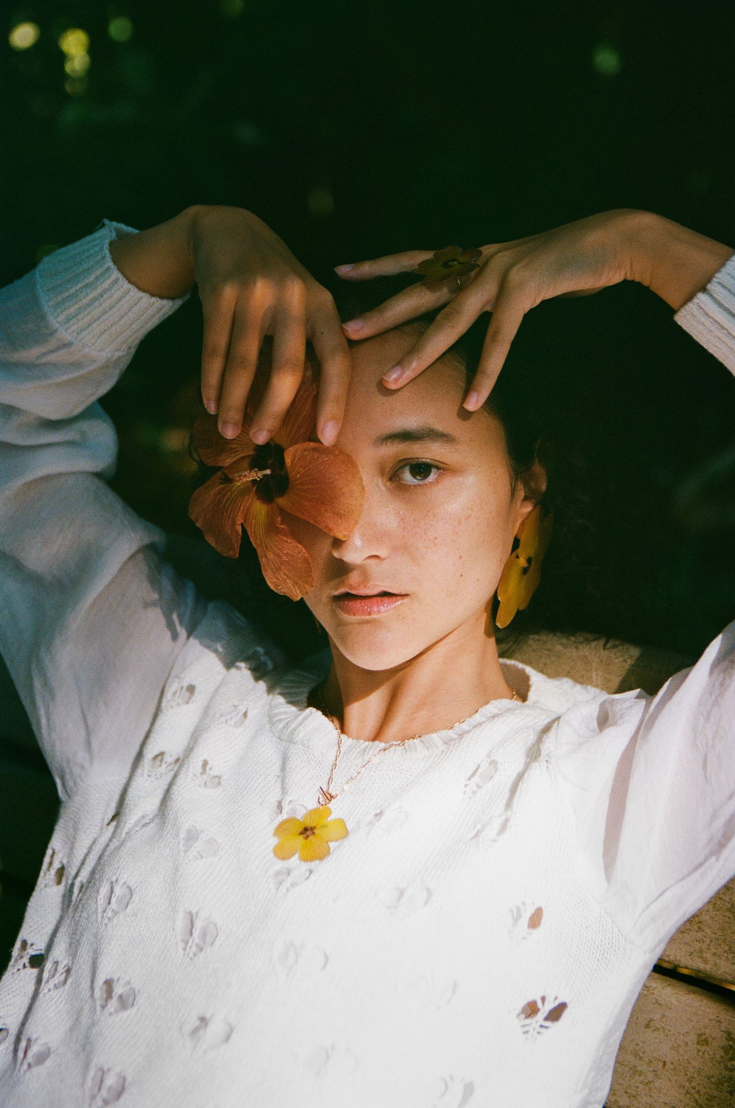 Woman in white floral top holding autumn leaf over face wearing yellow flower earrings and necklace.