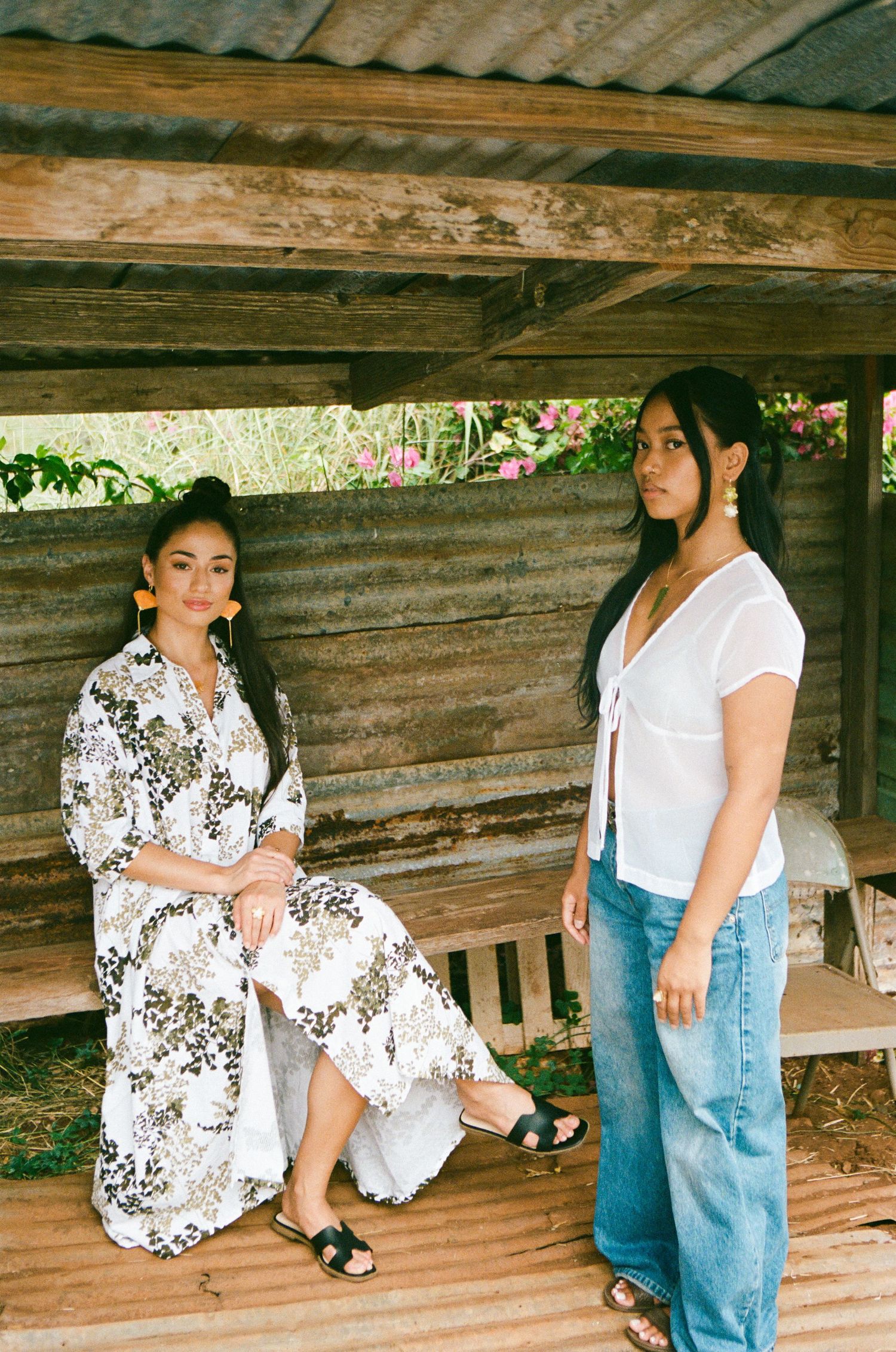 Two women posing under a rustic wooden shelter, one in a floral maxi dress and one in white top and jeans.