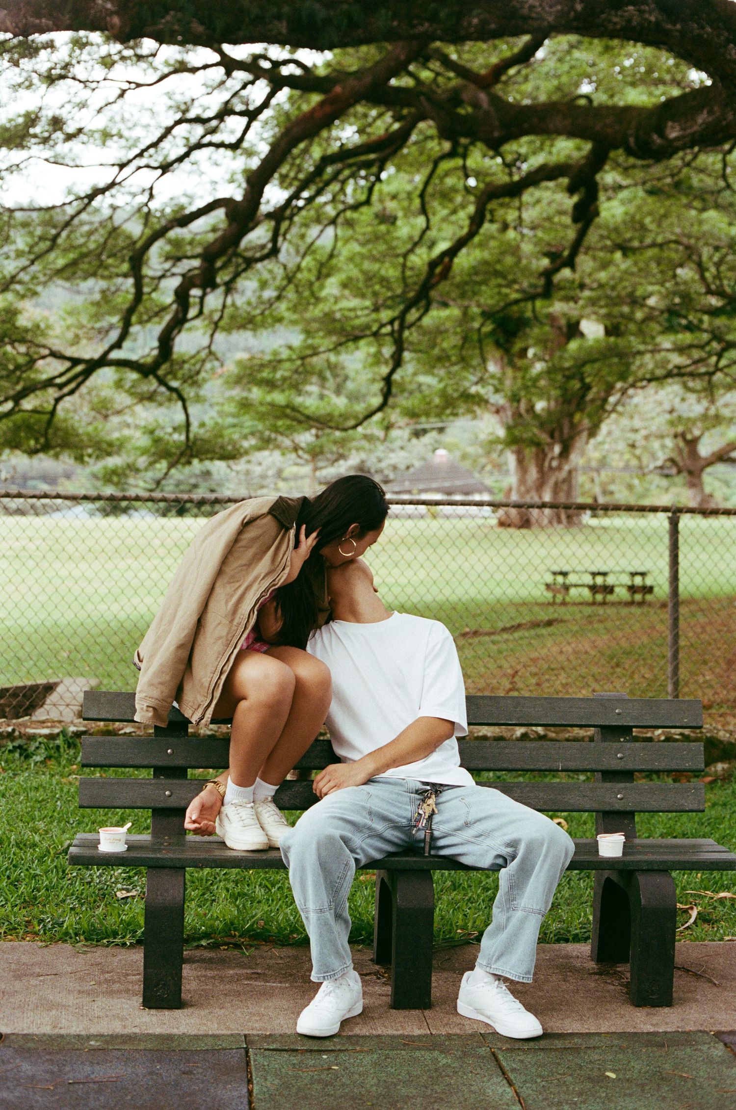 Couple sitting closely together on a park bench under large trees.