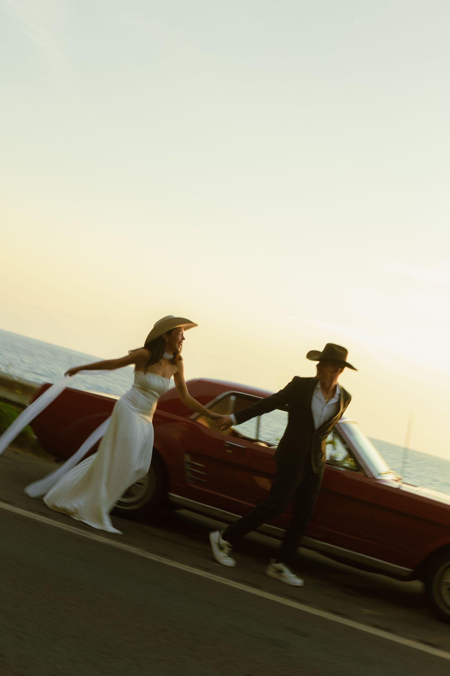 Bride and groom in cowboy hats posing beside a red vintage convertible at sunset.