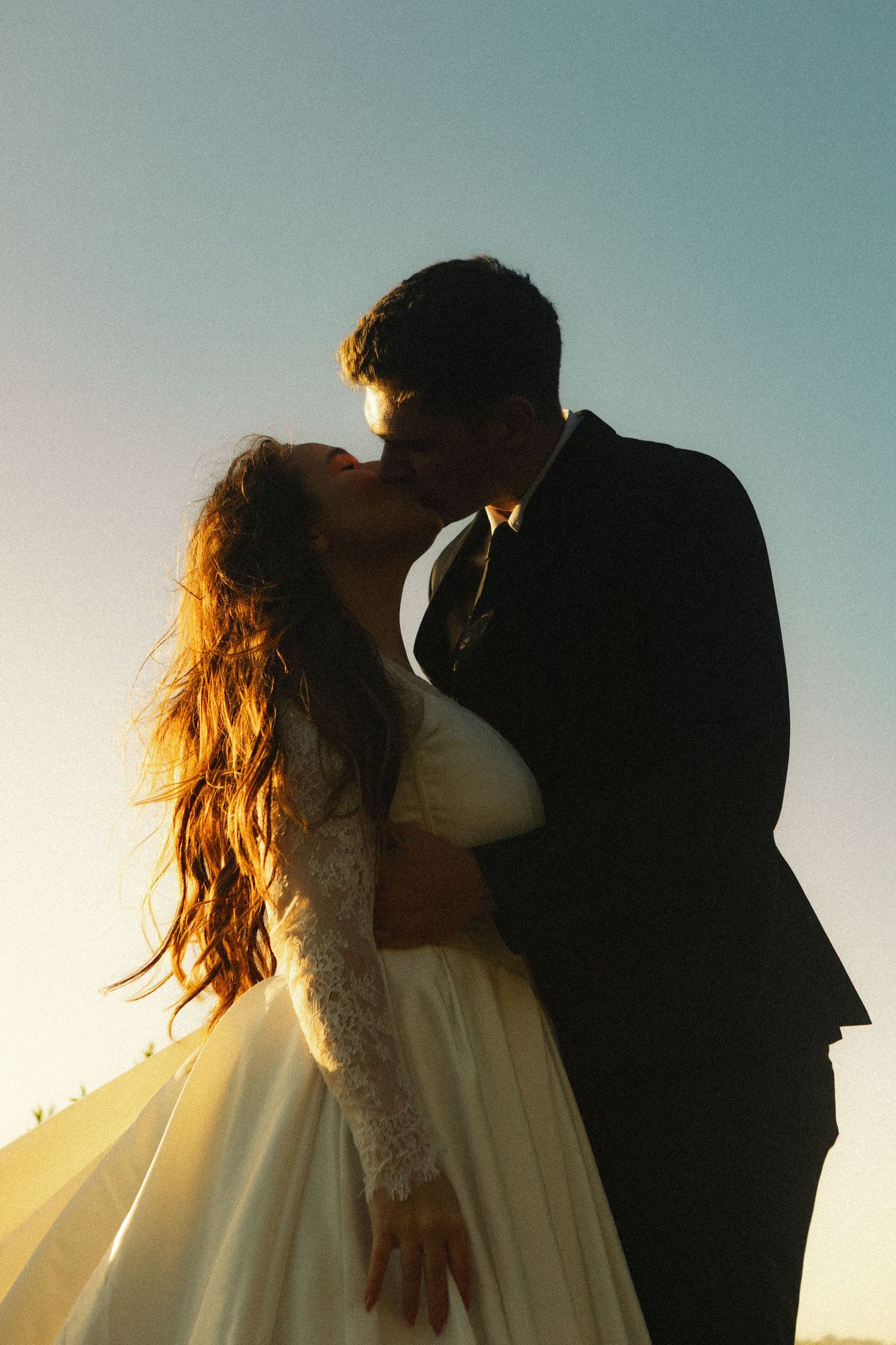 Bride and groom sharing a romantic sunset kiss, silhouetted against a clear sky, wedding dress and suit visible.