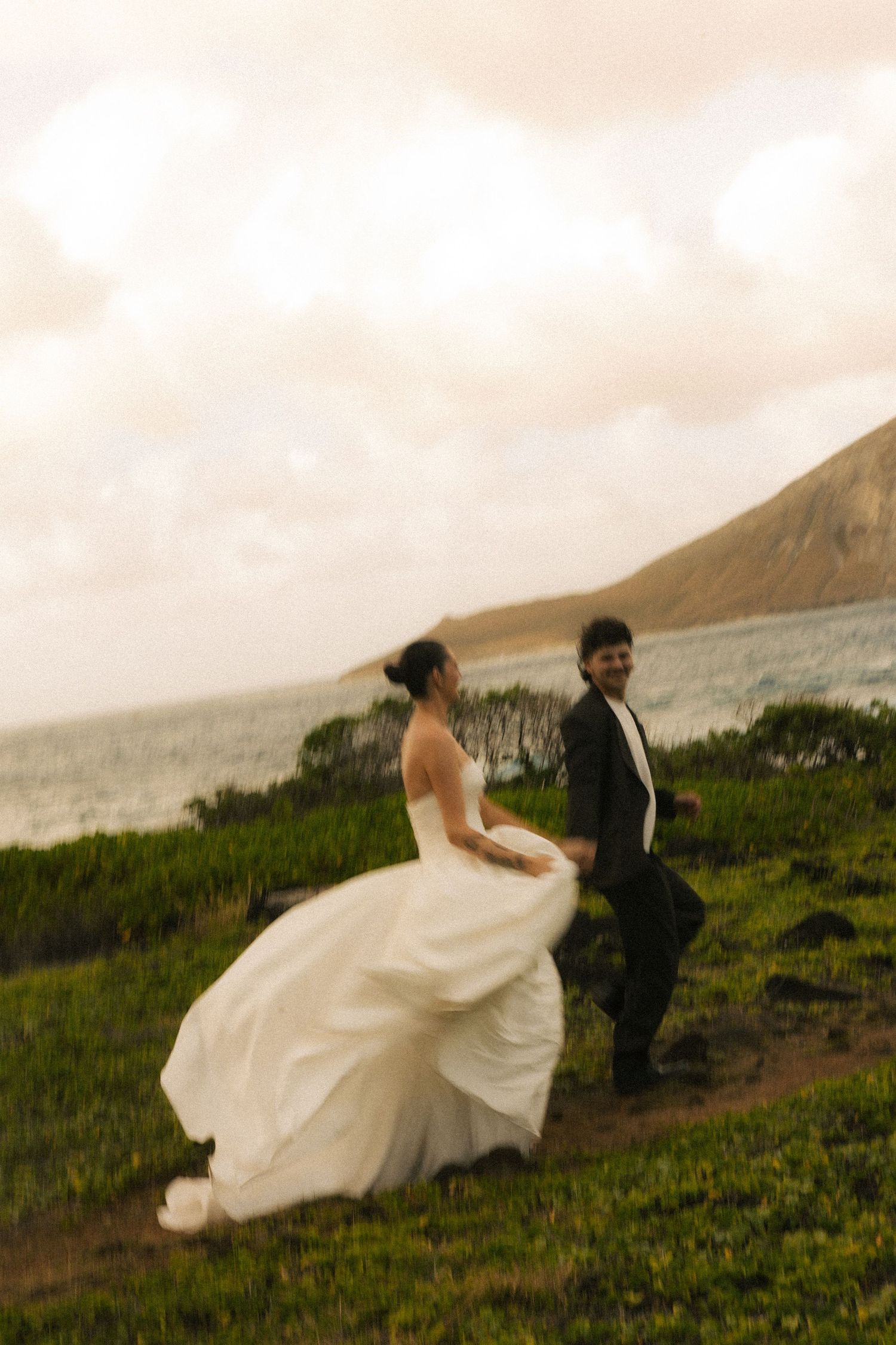Bride in flowing white gown and groom in black tux walk along coastal hillside with ocean backdrop.