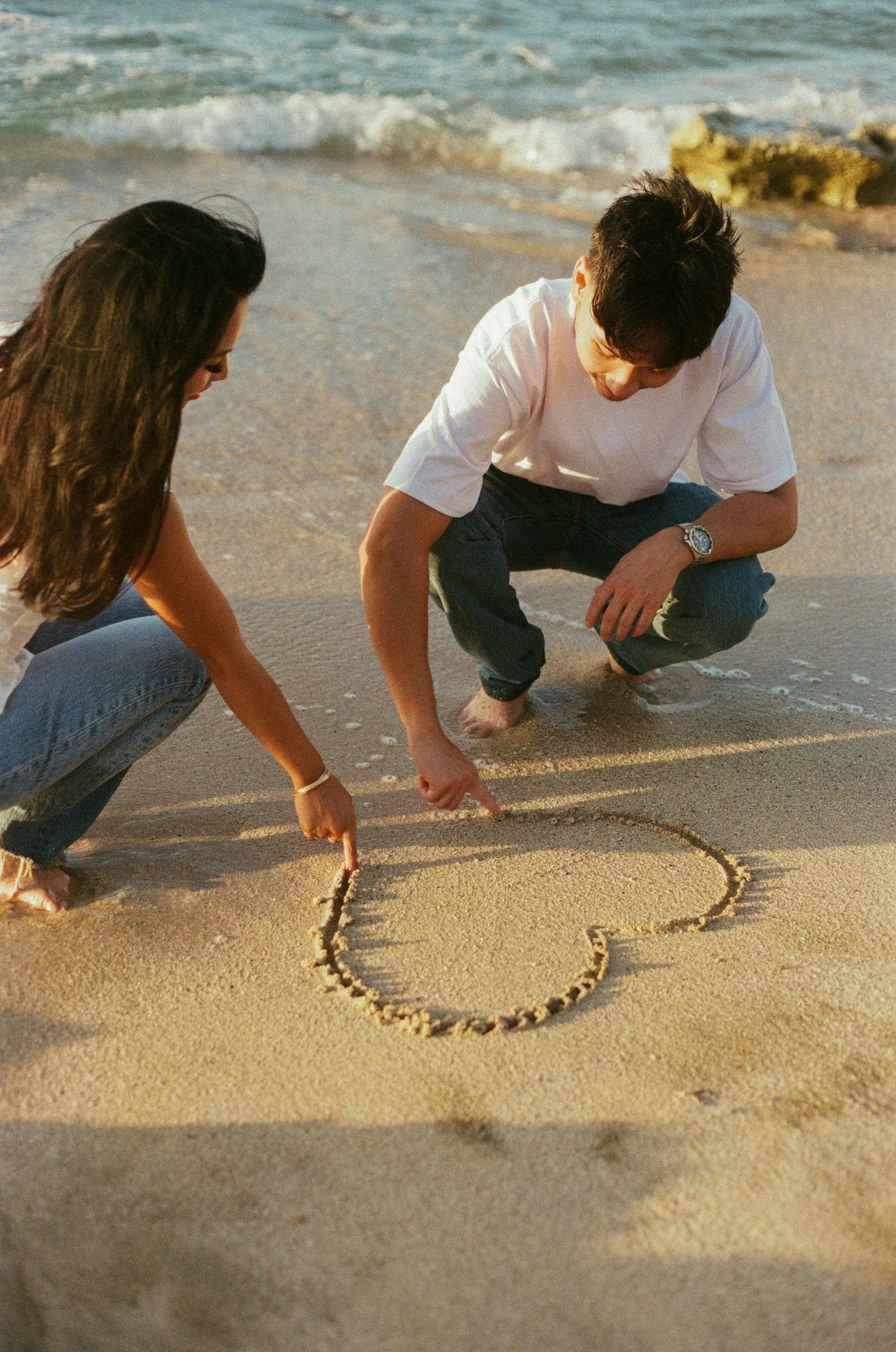 Couple drawing a heart shape together in wet sand at the beach.