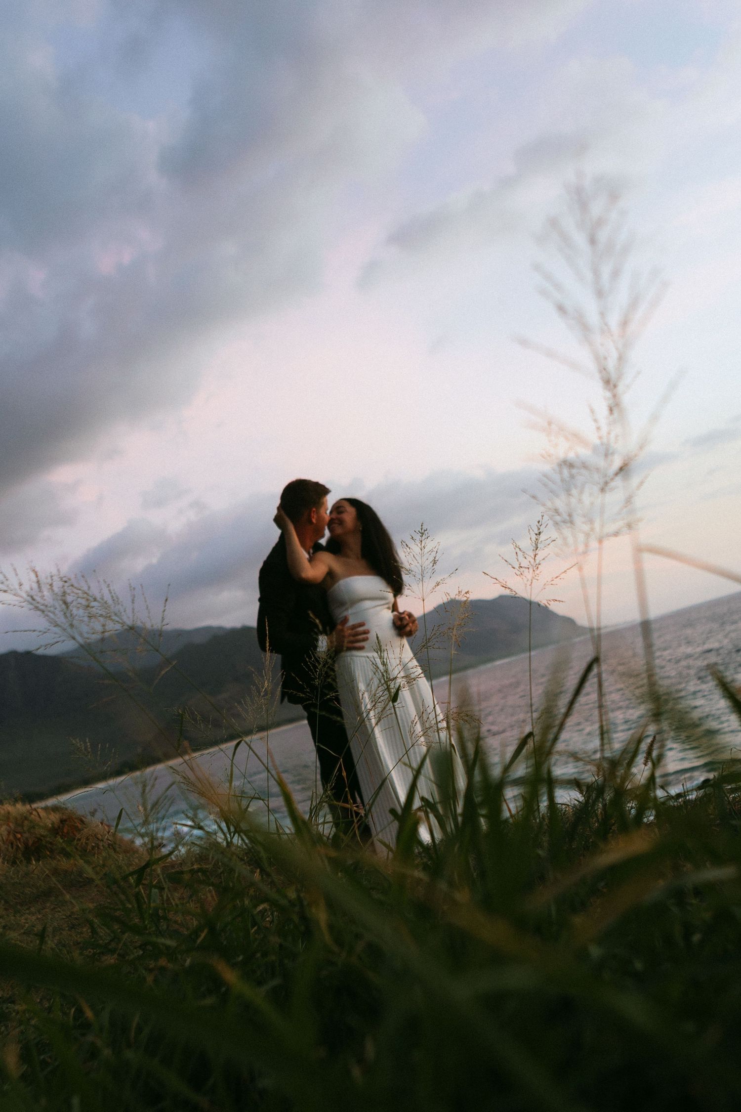 Couple kissing in white dress and dark suit on hillside with ocean and dramatic cloudy sky.