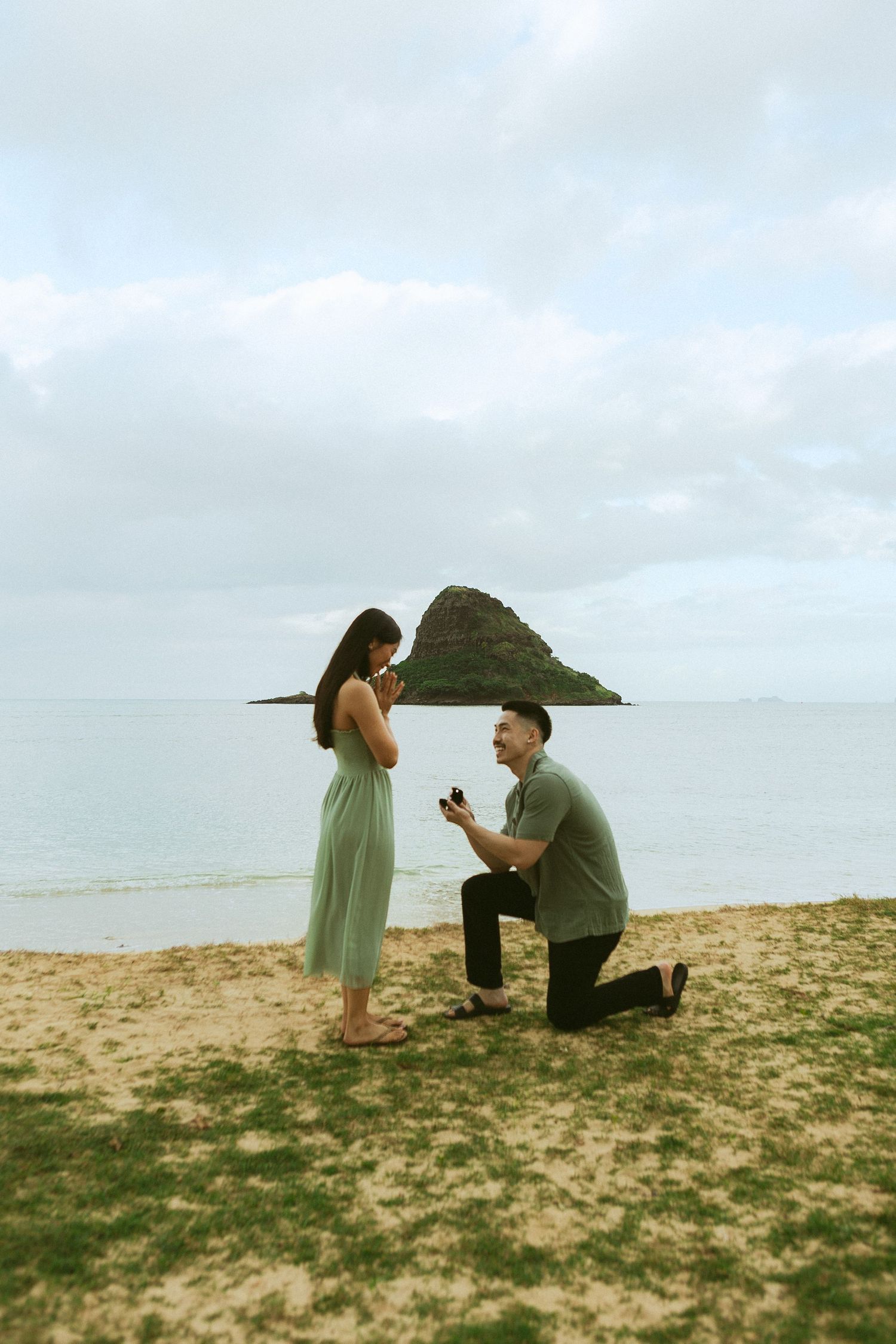 Man proposing on one knee to his girlfriend on a sandy beach with a small island backdrop.