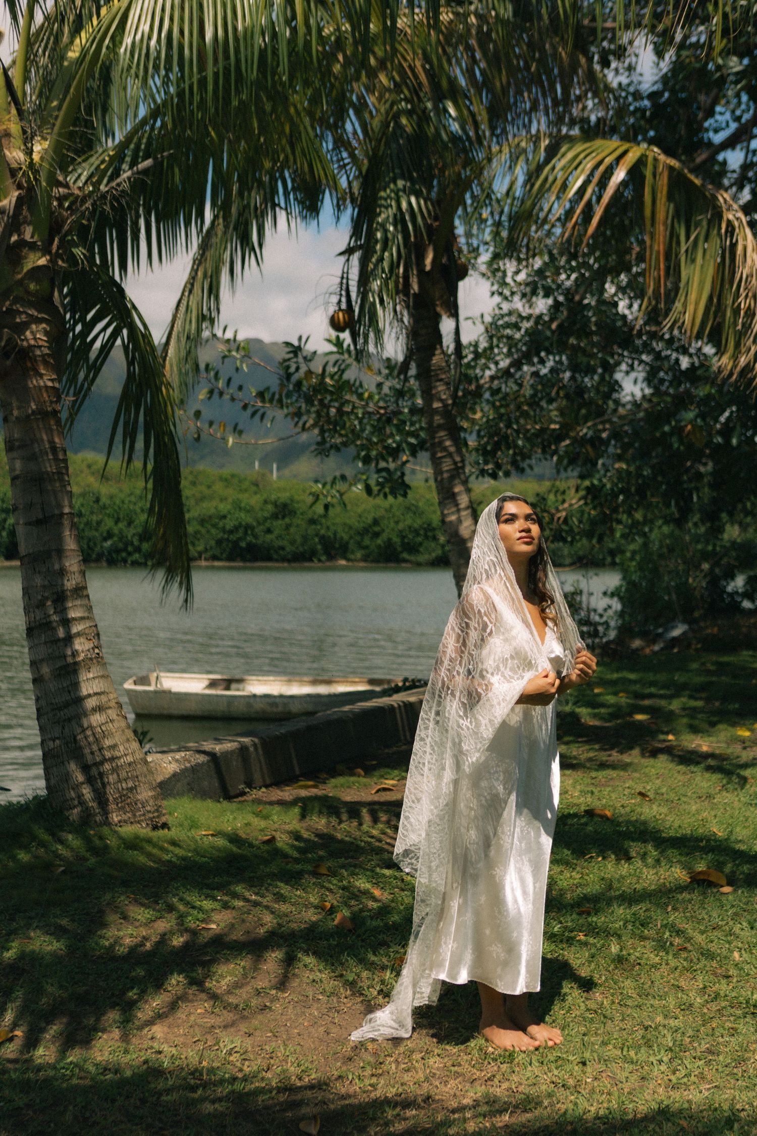 Bride in white lace veil and gown standing barefoot by tropical lake with palm trees and boat.