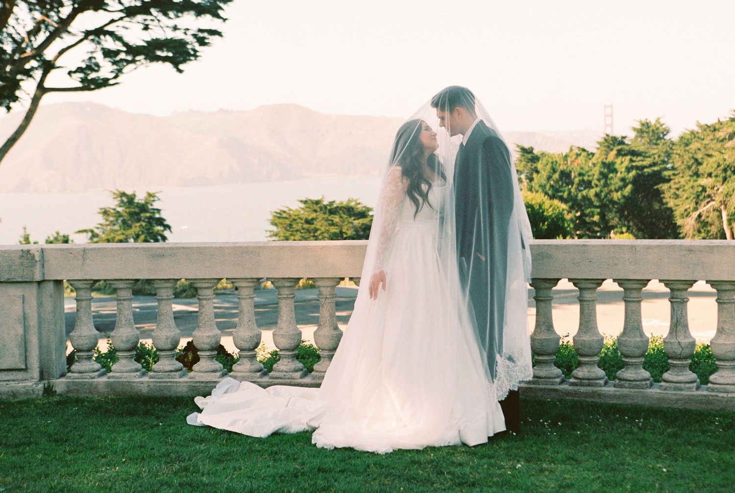 Bride and groom share a romantic kiss in front of a stone balustrade with mountain lake views on their wedding day.