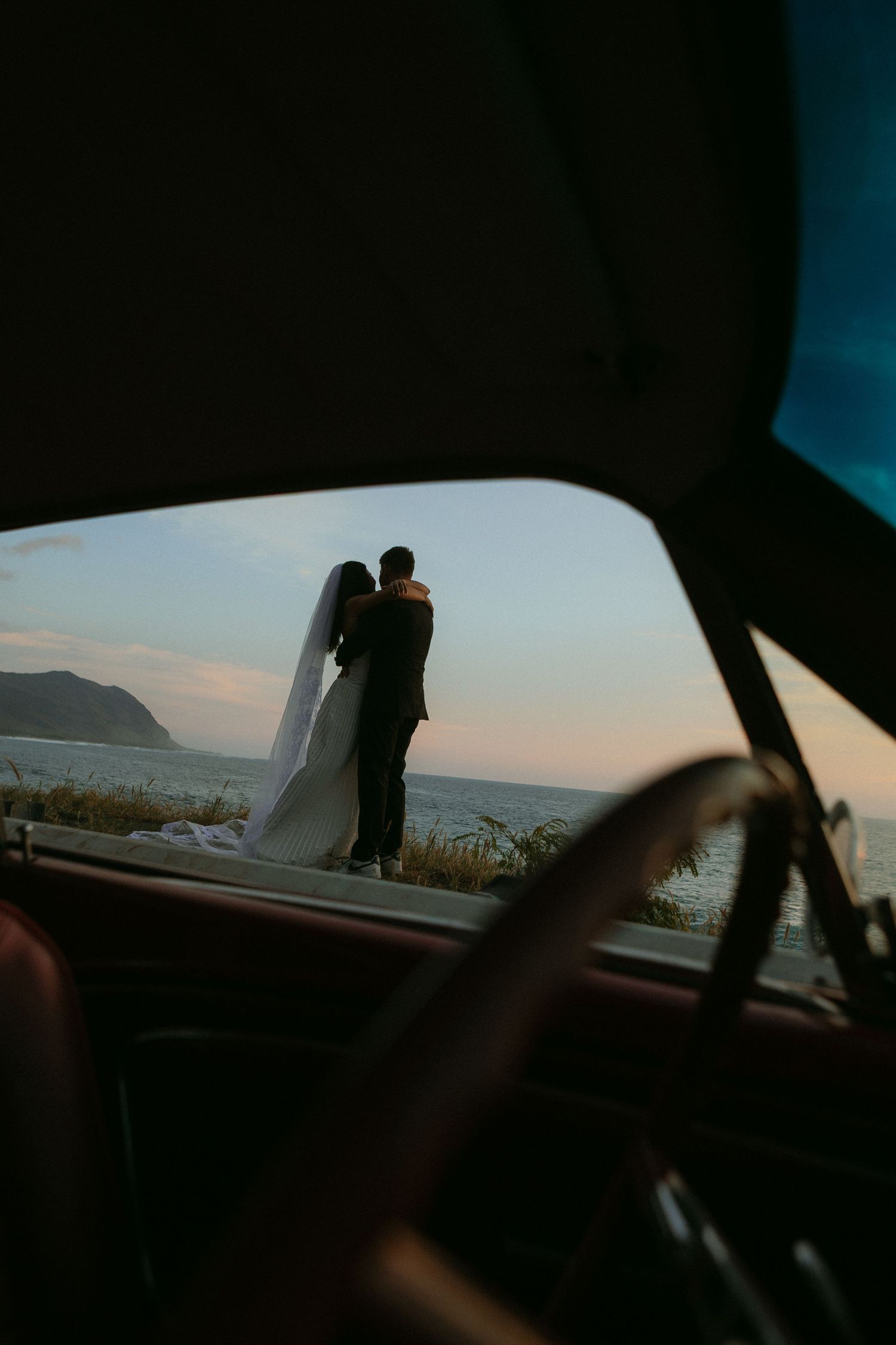 Bride and groom silhouetted at sunset by the ocean, viewed through a vintage car window.