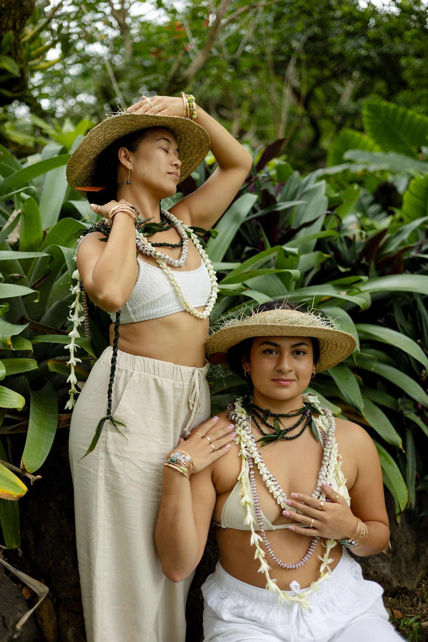 Two women in straw hats and white outfits wearing floral leis in a tropical garden.