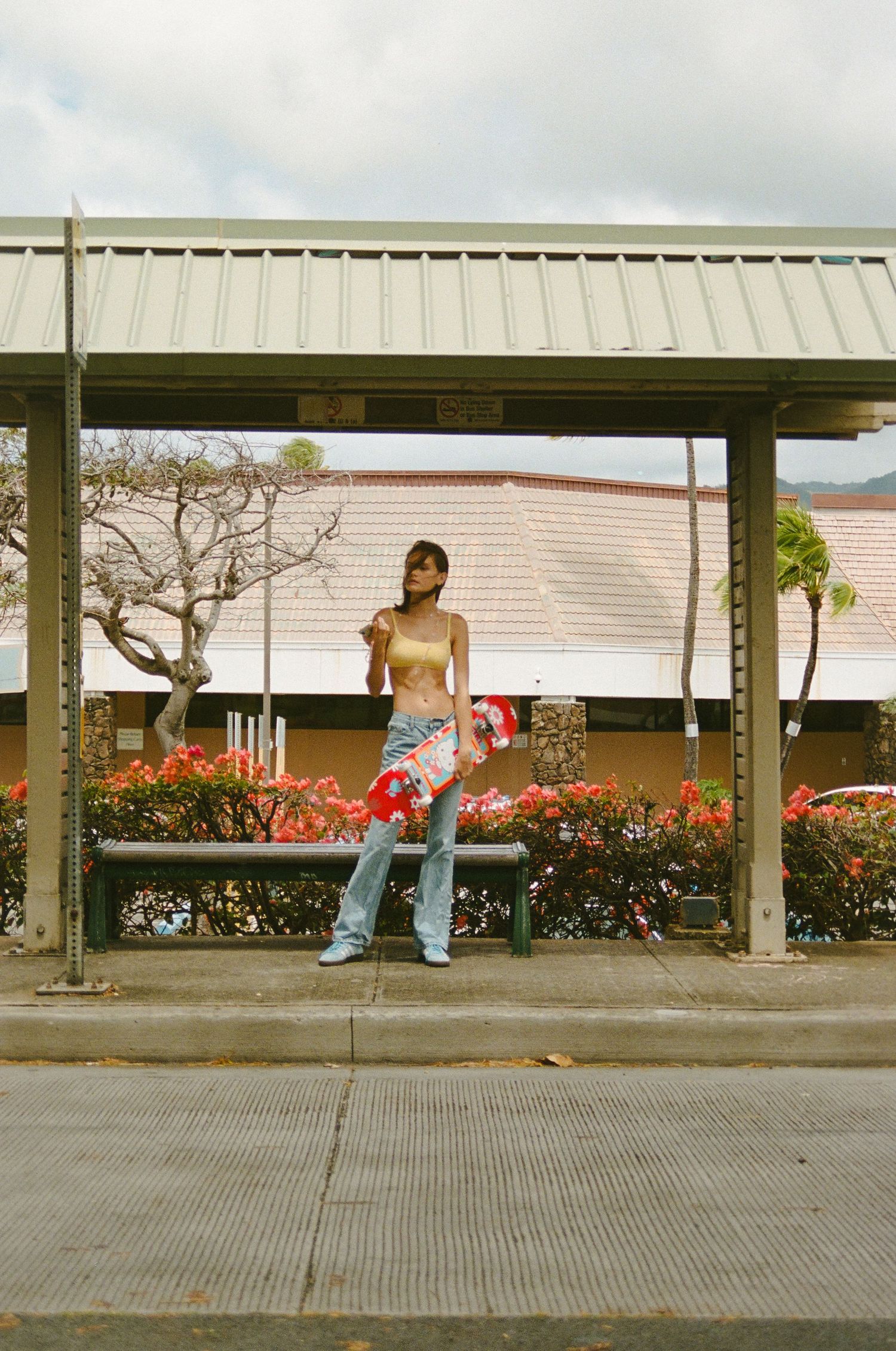 Woman in crop top and floral pants standing at a bus stop with red flowers behind her.