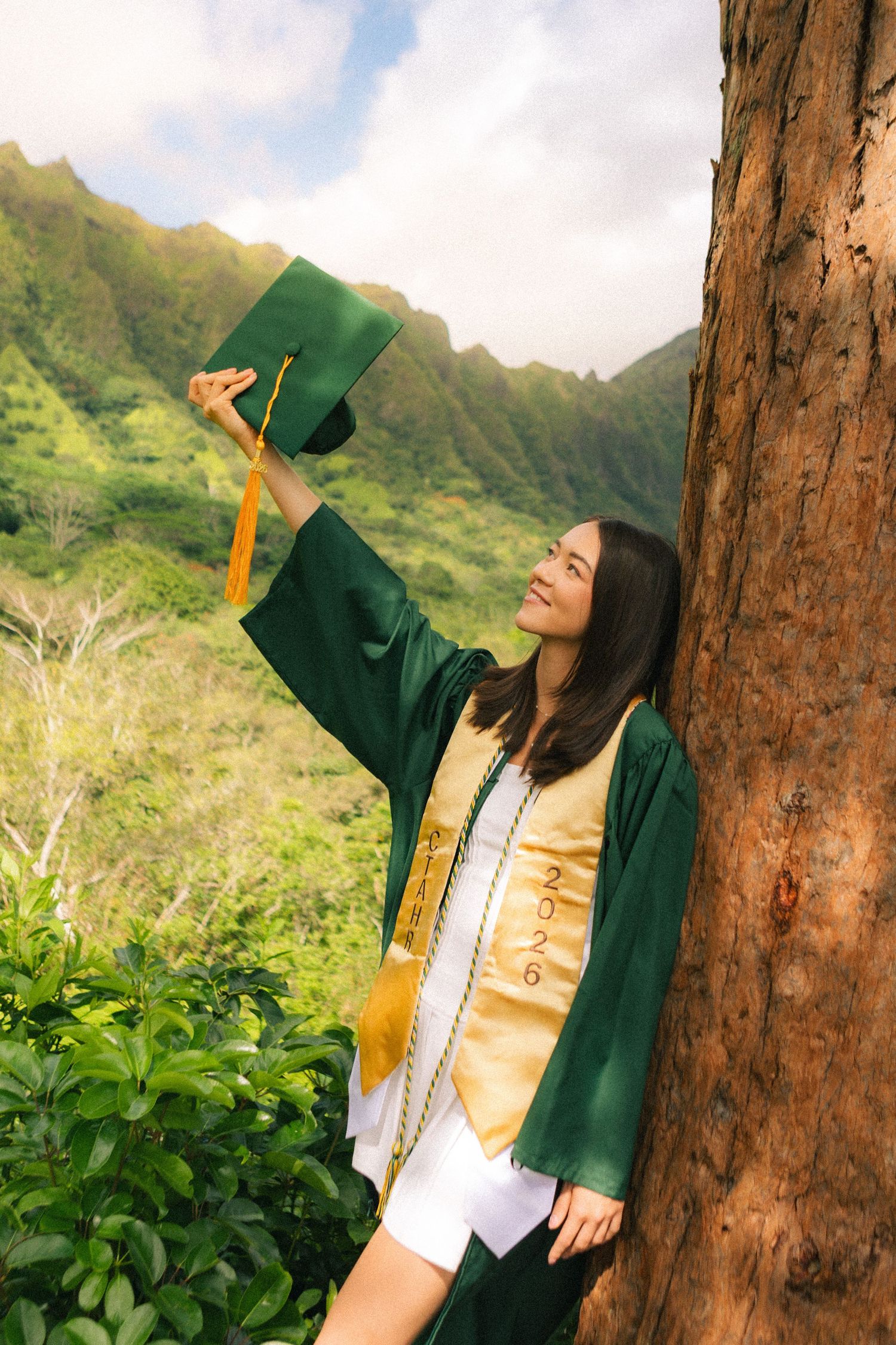 Graduate in green gown raises cap beside tree with scenic mountain backdrop in Hawaii.