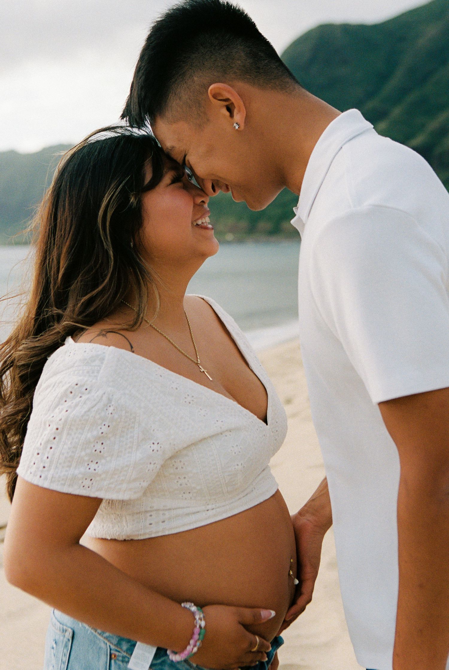 Pregnant couple sharing a tender moment on a sandy beach, wearing white outfits with mountains in the background.