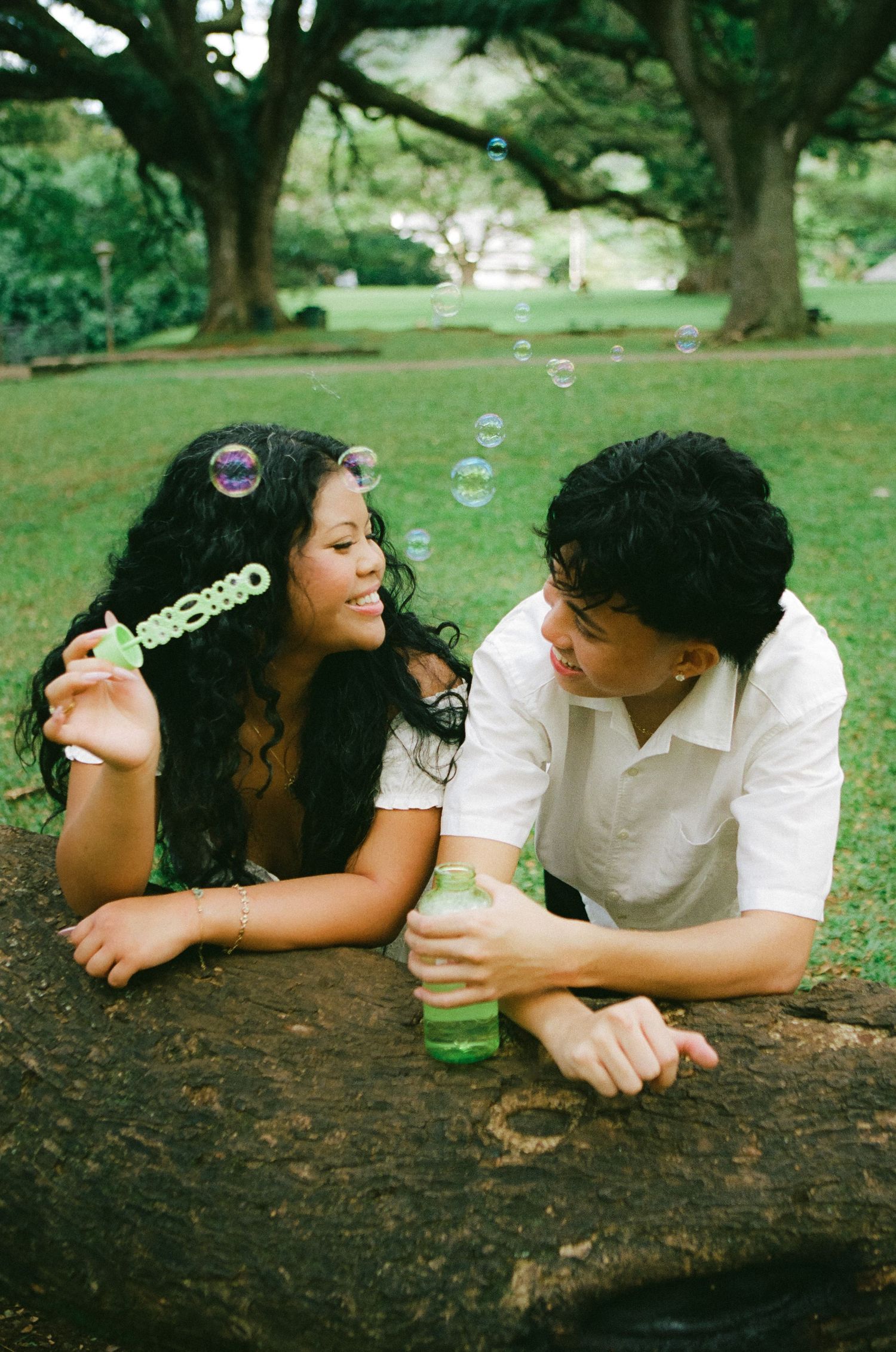 Young couple lying on rock blowing bubbles together in sunny park with large trees.
